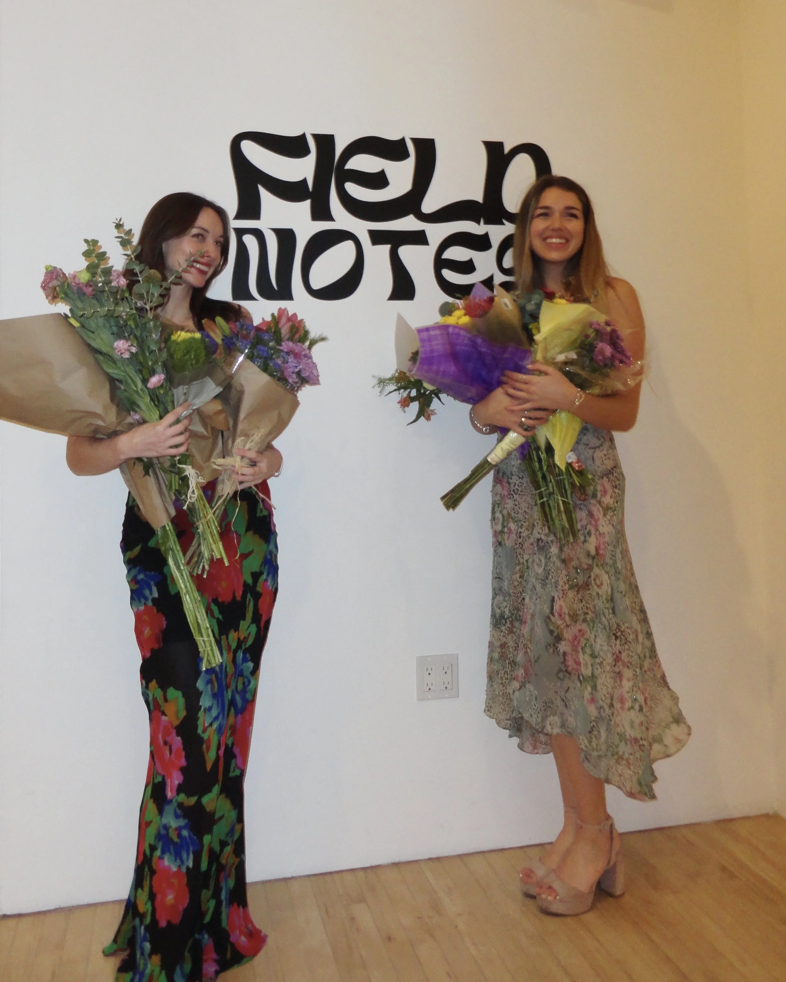 Two women standing side by side, each holding bouquets of flowers, smiling, with a white wall and black text in the background.