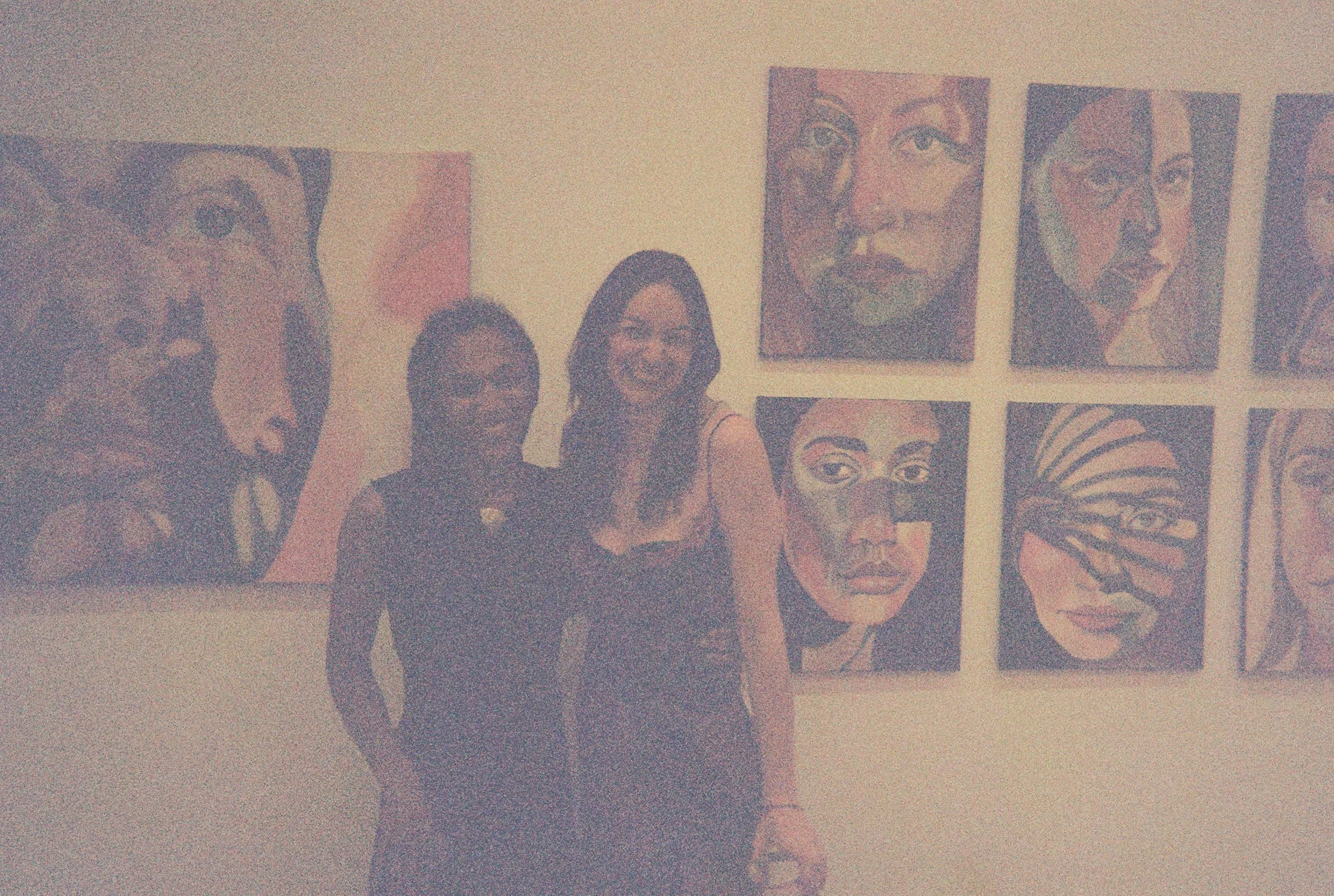 Two women standing in front of a wall of colorful paintings of faces at an art gallery.