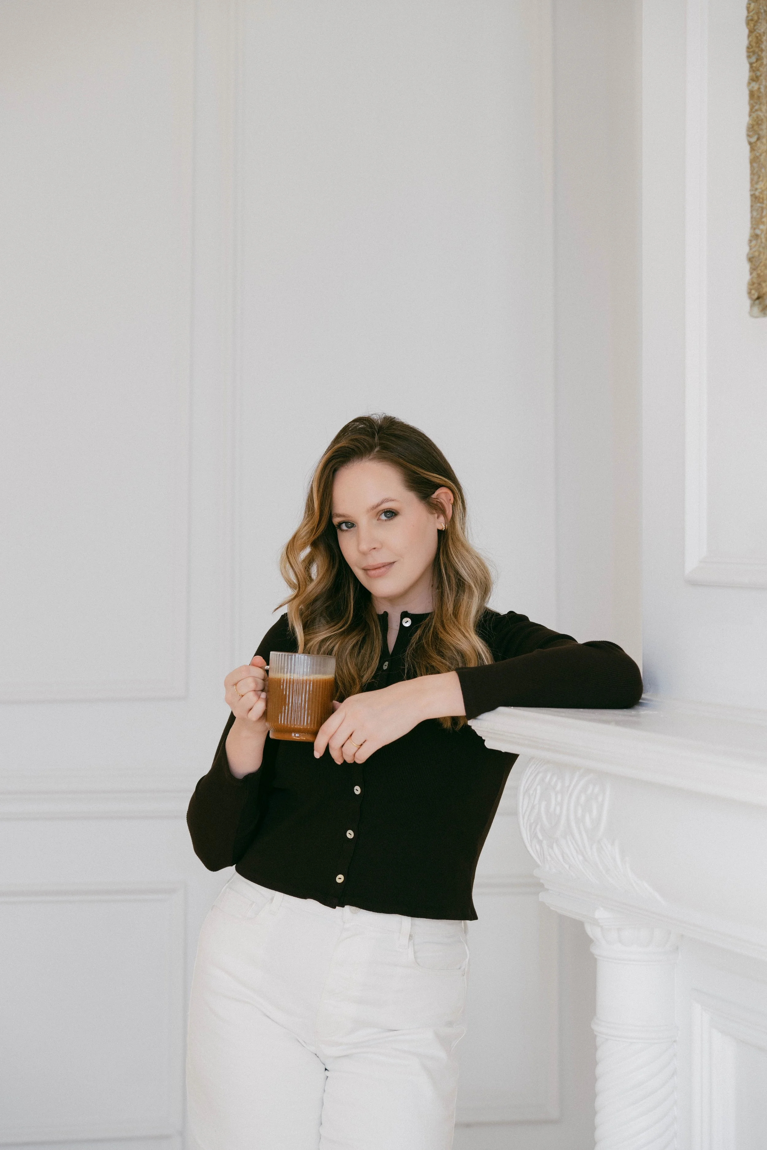Courtney Slate, a woman with wavy brown hair in a black top with white pants holding a mug, standing in a white ornate room, leaning on a decorative white fireplace.