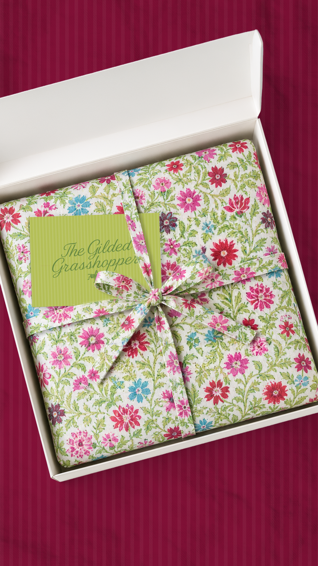Tablecloth gift packaging wrapped in pink and green floral fabric with a Gilded Grasshopper card, on a mulberry striped background