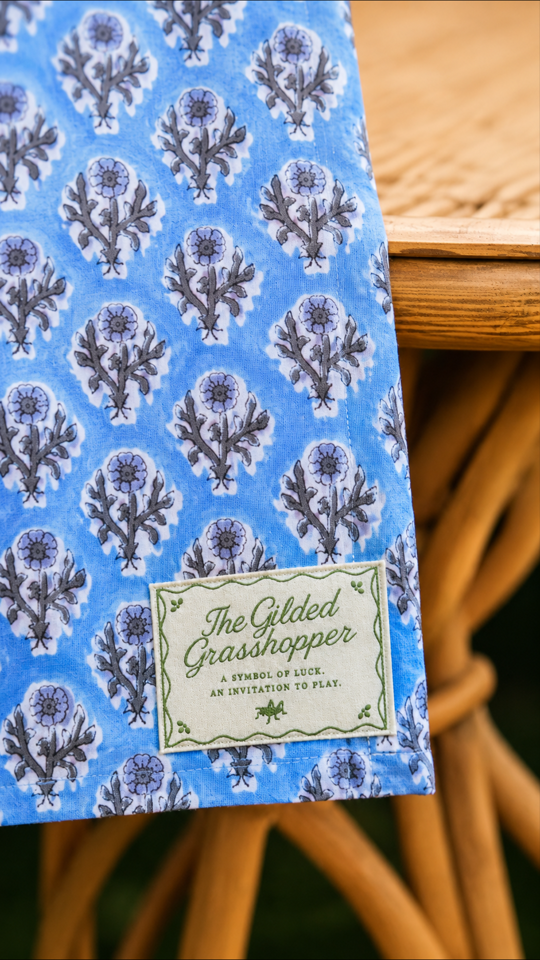 Blue floral block-print tablecloth with an embroidered Gilded Grasshopper label, draped over a rattan table