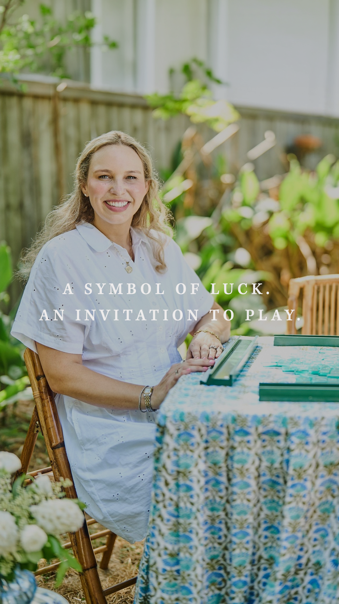 Founder Lauren Davis at a mahjong table set with a blue block-print tablecloth, with the tagline "A Symbol of Luck. An Invitation to Play."