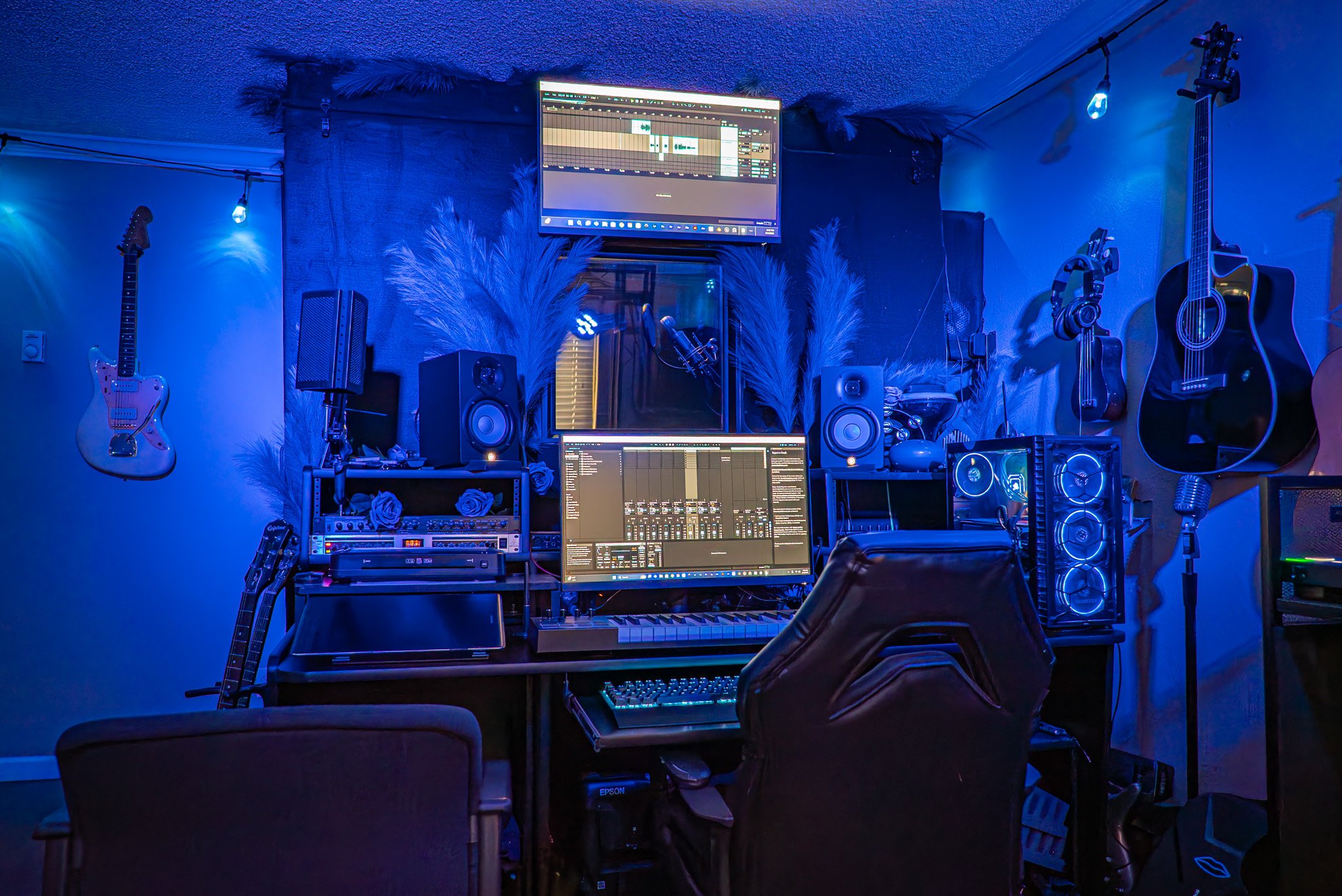 A music production studio with blue lighting, featuring multiple guitars hanging on the wall, speakers, a computer with a digital audio workstation on the screen, a keyboard, and other audio equipment.