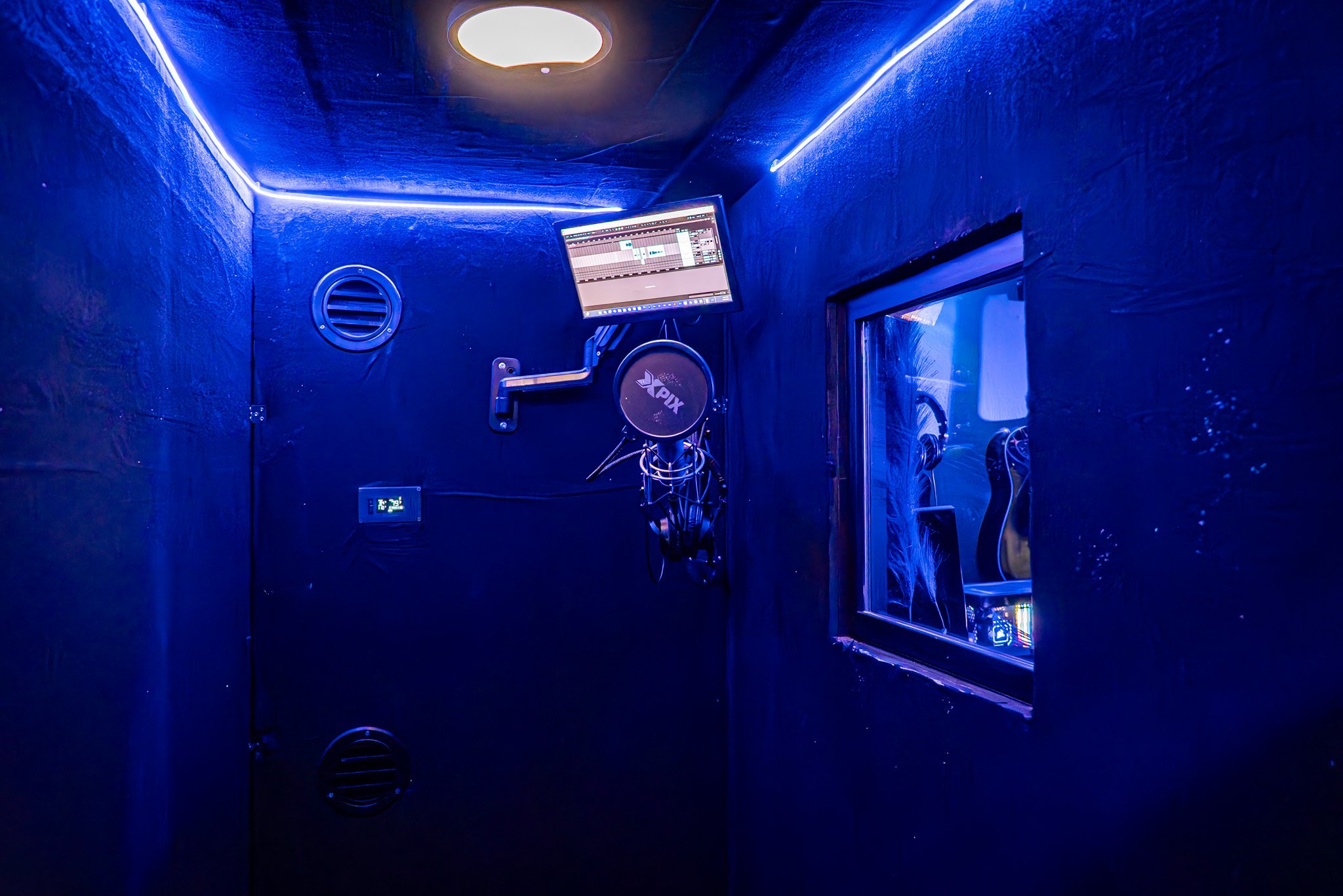 Soundproof recording booth with blue walls, a hanging microphone, a computer monitor, a window showing a sound engineer's workspace, and musical instruments visible through the window.