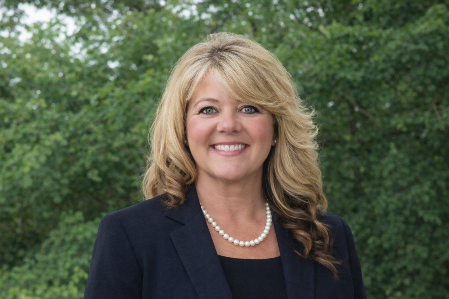 Woman with blonde hair smiling, wearing a black off-shoulder top and gold necklace, standing outdoors with green trees in the background.