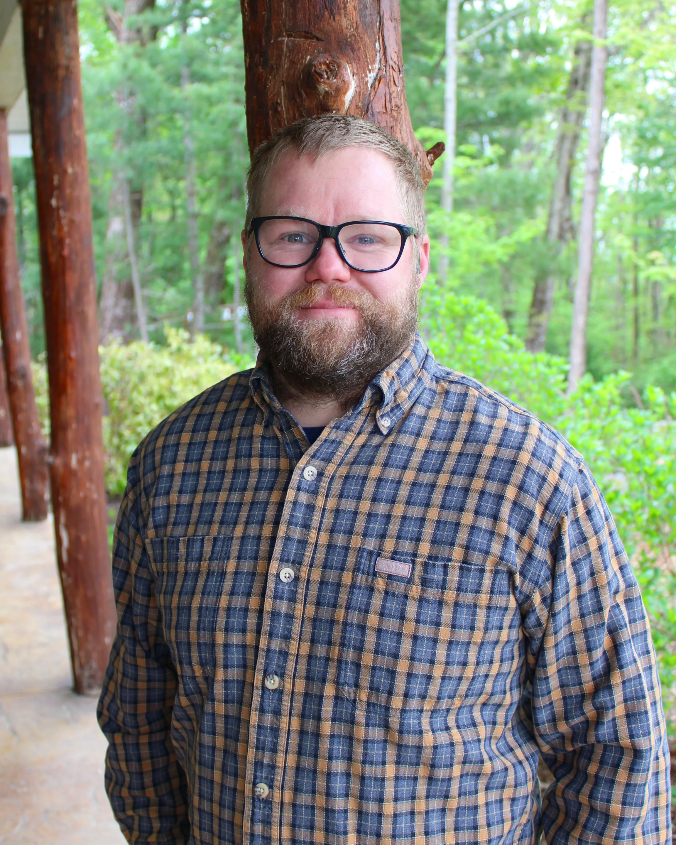 Man with glasses wearing a plaid shirt and a maroon shirt standing on a porch with greenery in the background.