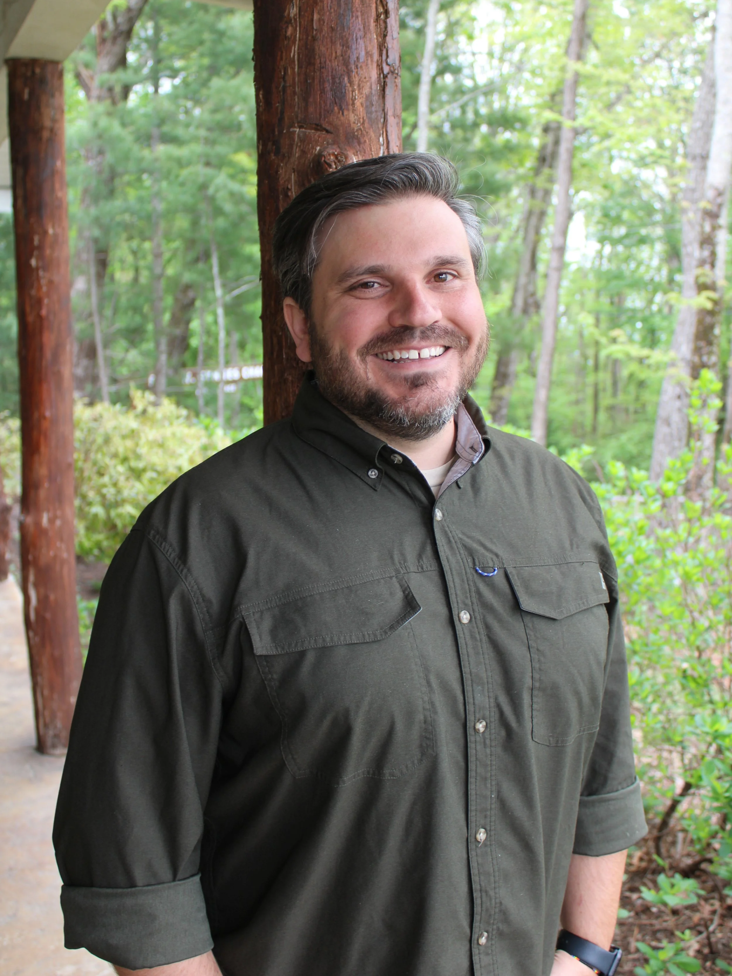 Man with beard in dark green shirt standing outdoors next to trees.