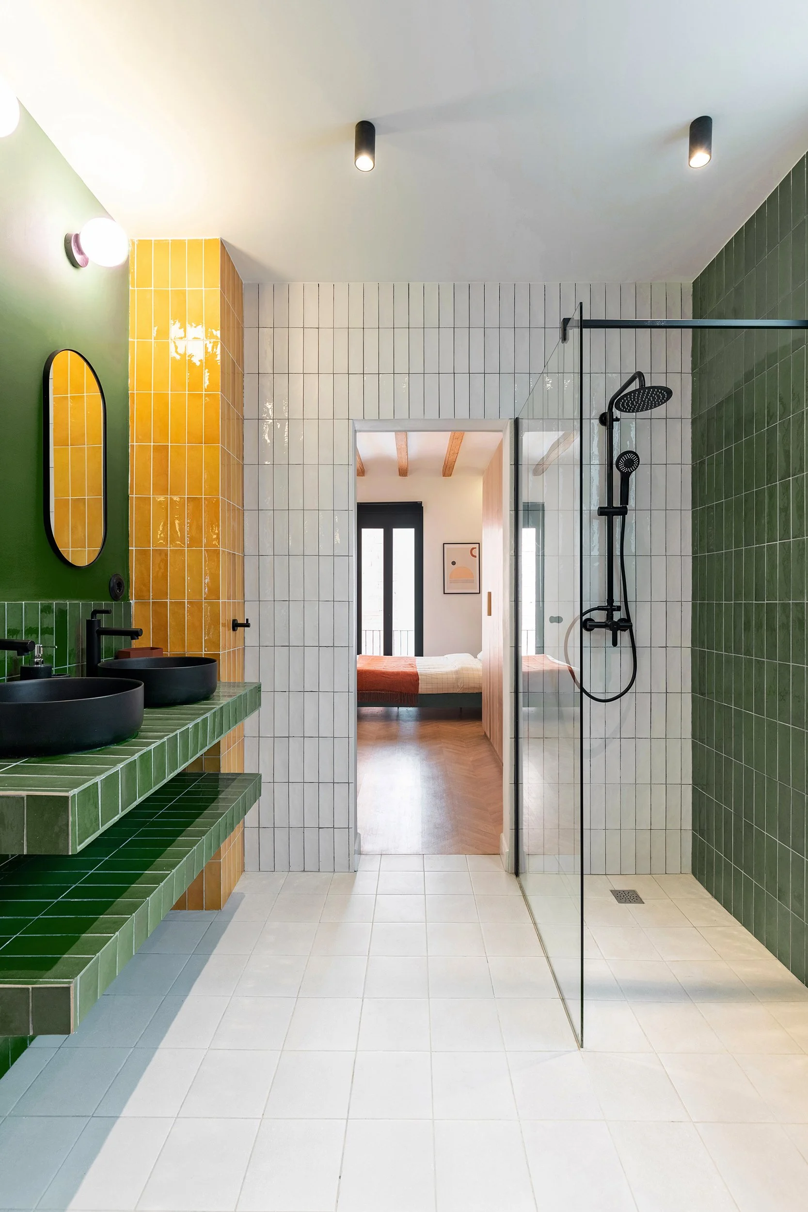 Modern bathroom with green, yellow, and white tiles, featuring a glass shower, black fixtures, and an adjacent bedroom doorway.
