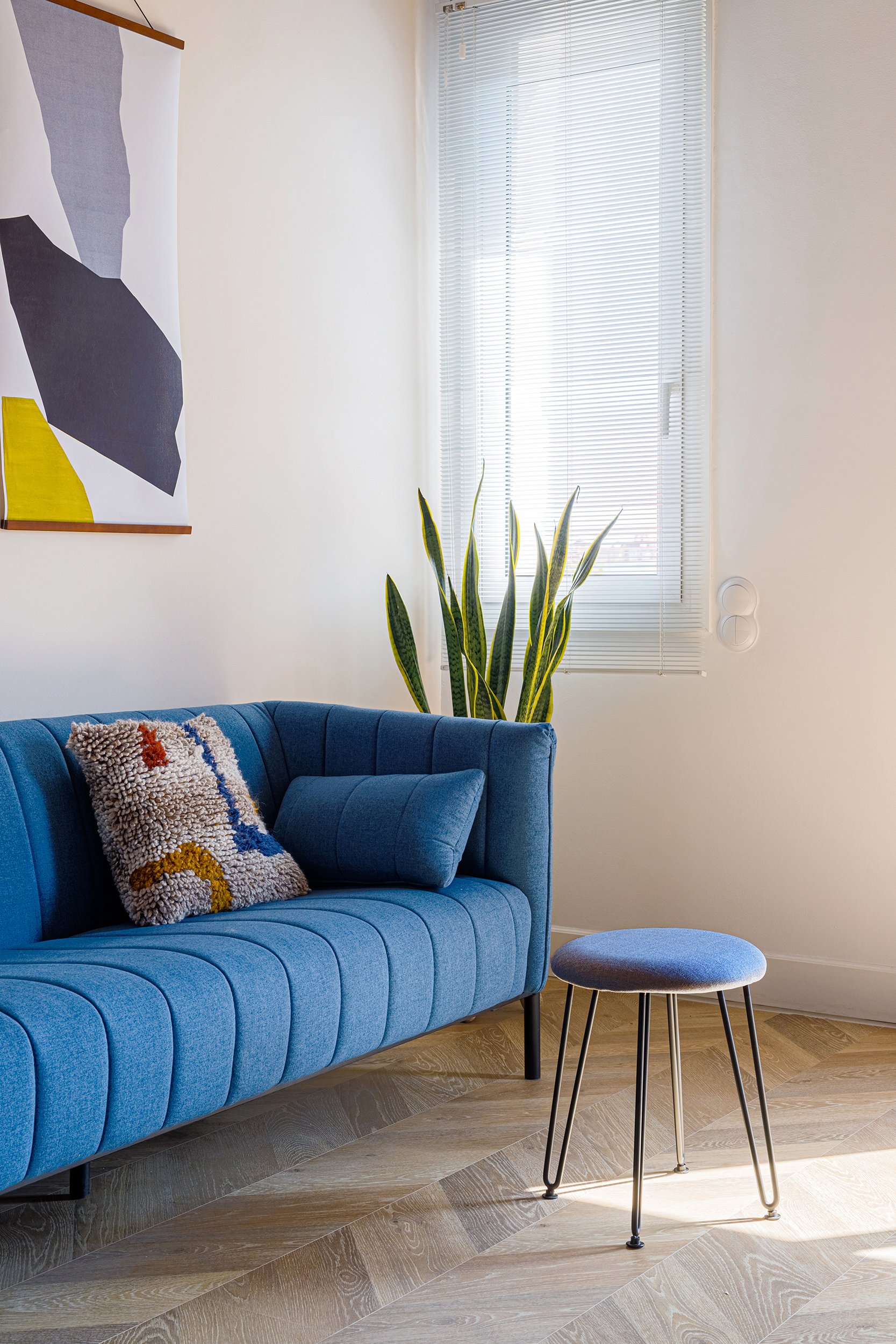 Living room with a blue sofa, textured pillow, small blue stool, abstract wall art, potted plant, window blinds, and wooden floor.