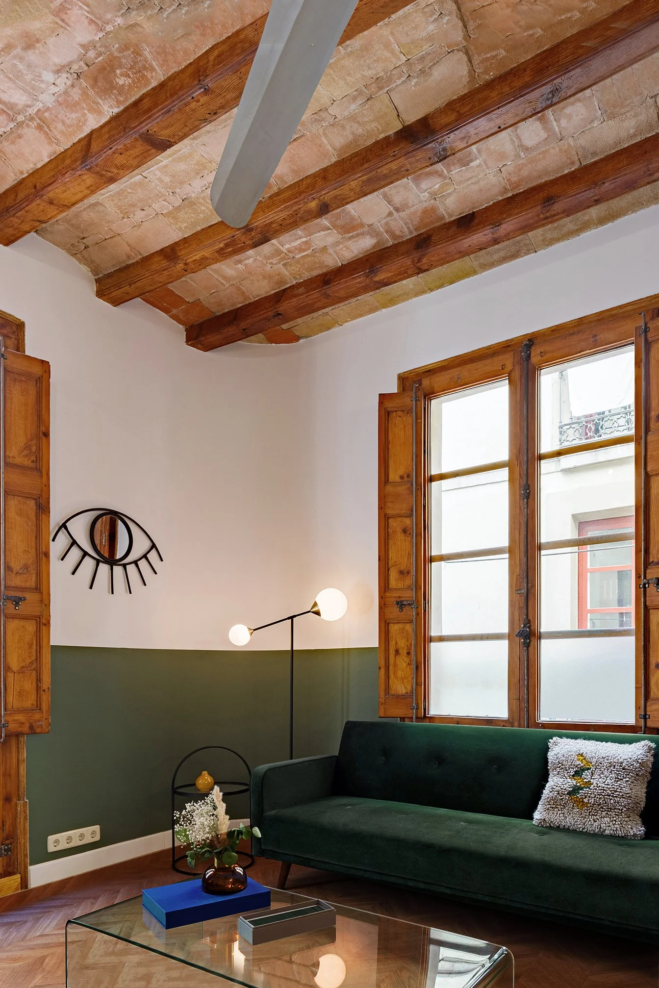 Cozy living room with exposed wooden beams, green velvet sofa, decorative pillow, eye-shaped wall art, floor lamp, large windows, and glass coffee table.