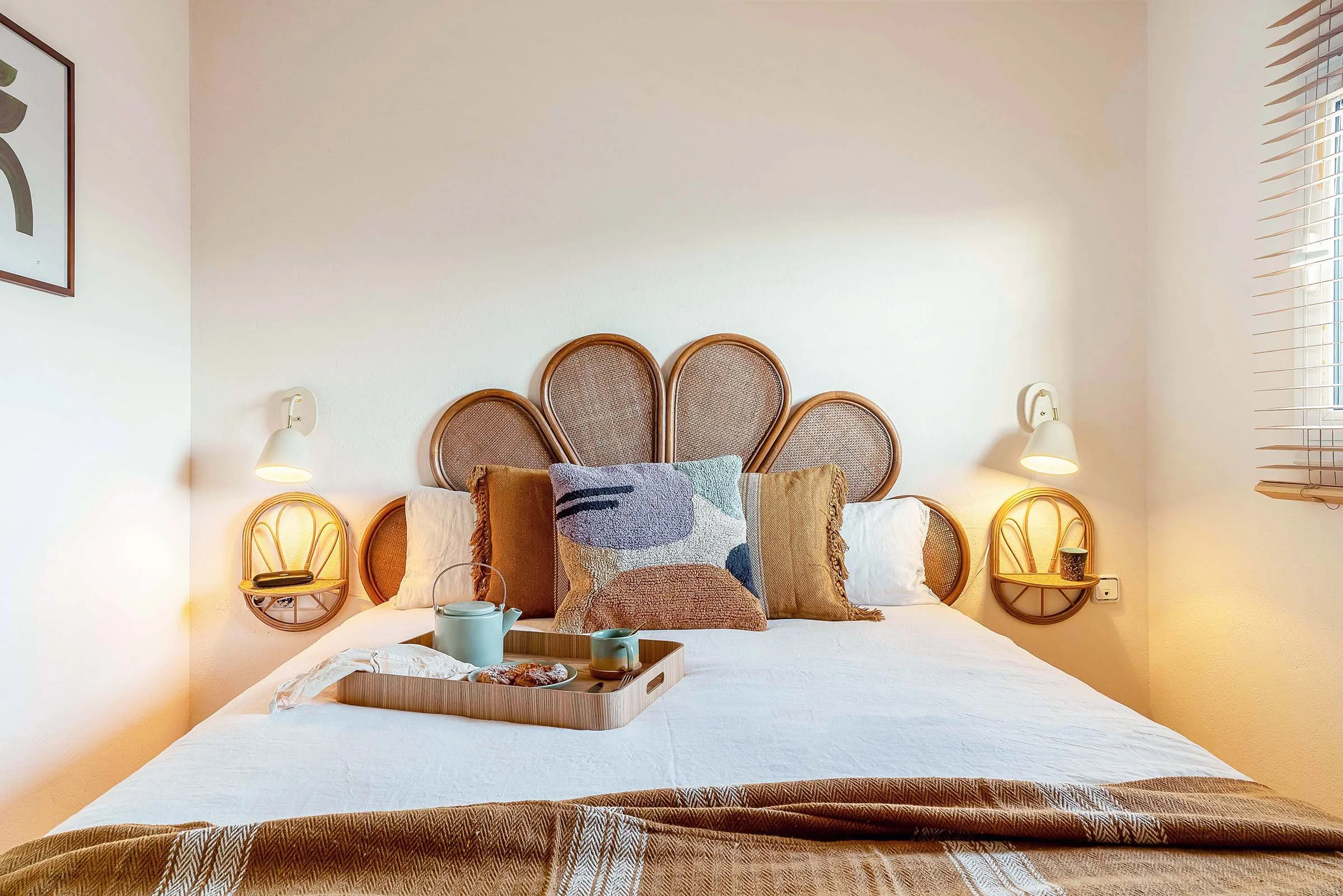 Cozy bedroom interior with a rattan headboard and a neatly made bed. The bed features white and brown pillows and a wooden tray with tea and pastries. Two wall lamps are above small shelves beside the bed, and a window with blinds is on the right.