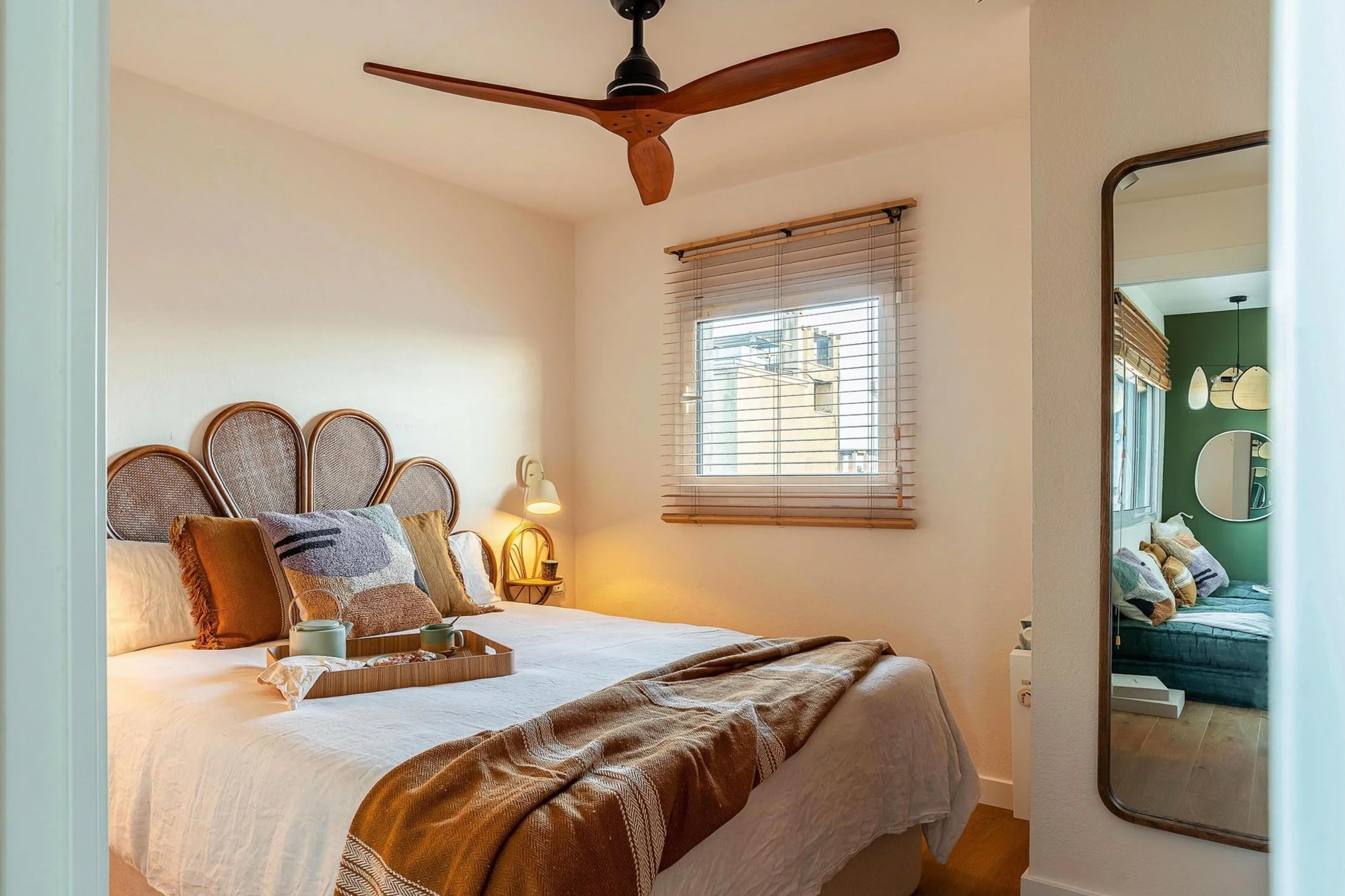 Cozy bedroom with a bed featuring a wicker headboard, pillows, and a brown throw. A wooden tray with a teapot and cups rests on the bed. A window with blinds allows natural light. A reflection in a wall mirror shows another part of the room with a gr