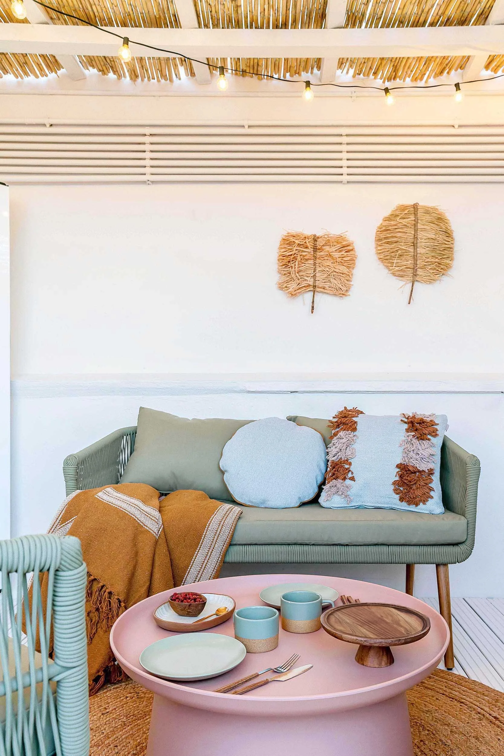 Cozy sitting area with a sage green sofa, decorative pillows, and a mustard throw blanket. A round pink coffee table with mugs, plates, and cutlery. Woven wall decor and string lights on a wooden ceiling.