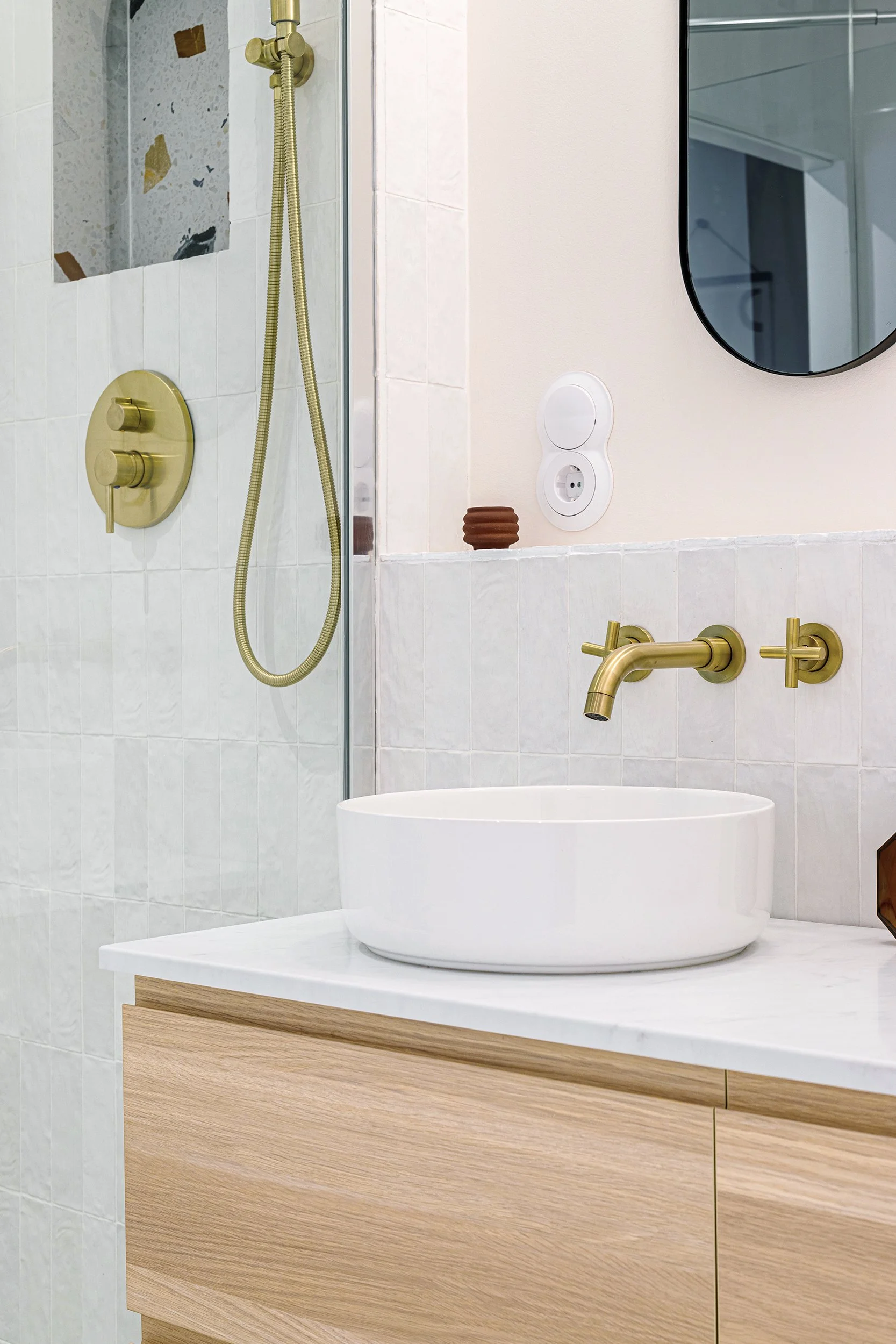 Modern bathroom with round white vessel sink, brass faucet, and shower fixture. Light wood vanity with marble countertop and mirrored wall.