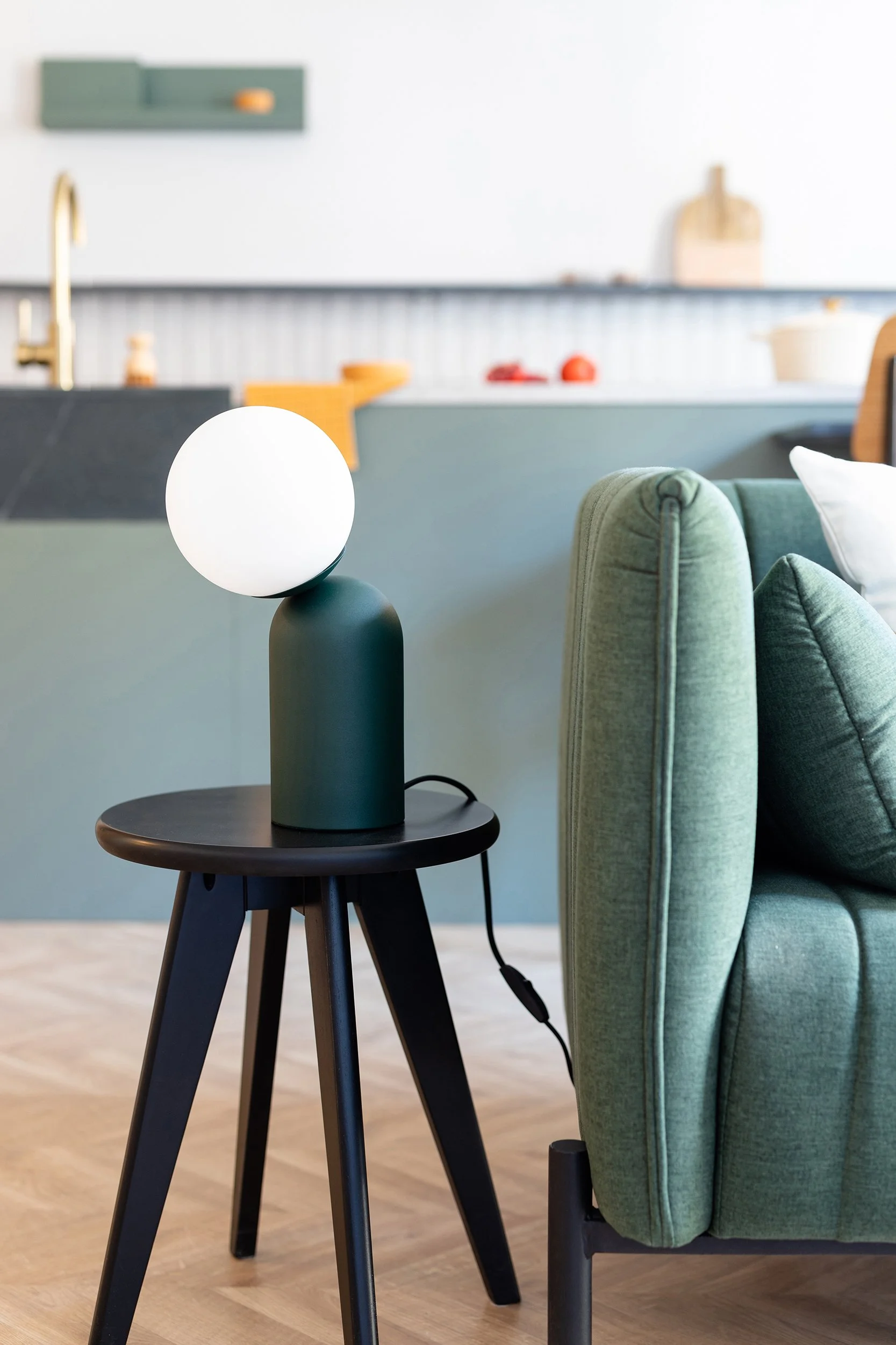 Modern living room with a green sofa and a small black side table featuring a contemporary lamp. Background includes a kitchen counter with a gold-colored faucet and blurred kitchen items.