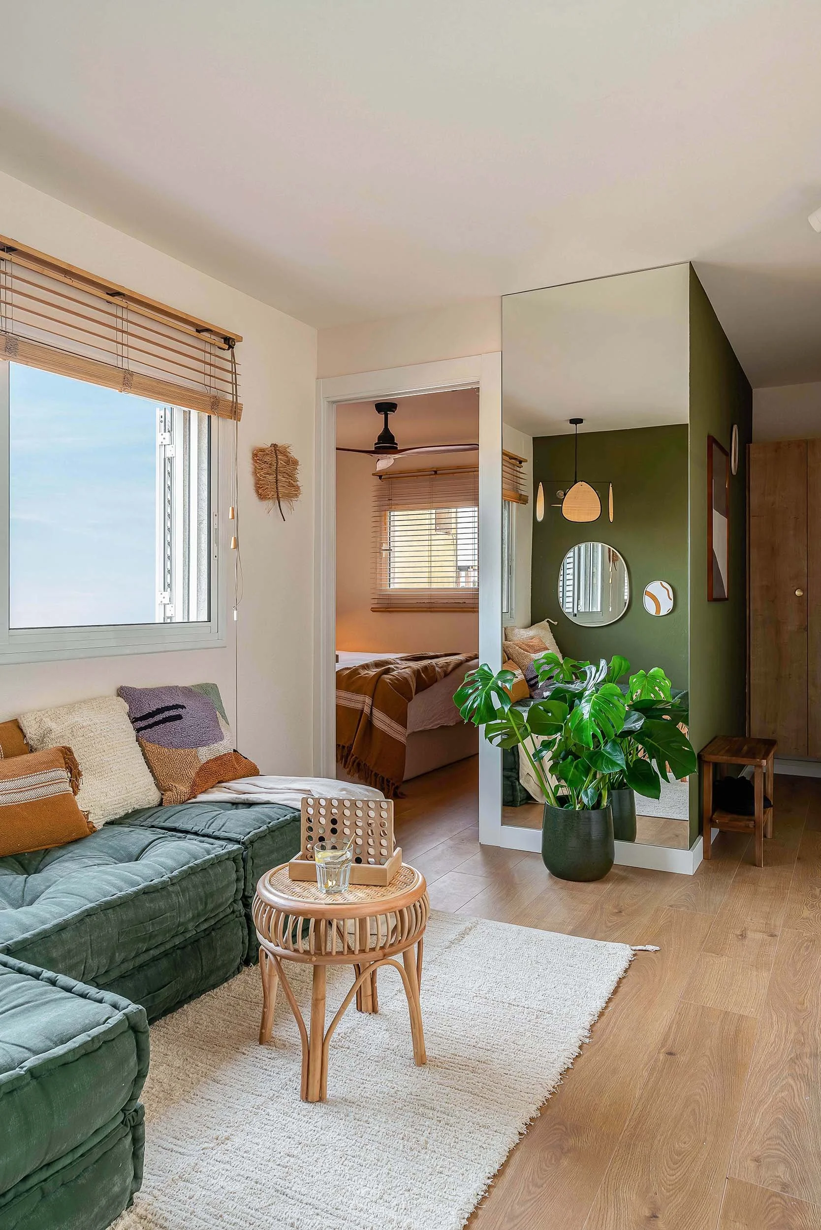 Modern living room with green sectional sofa, rattan side table, and indoor plant. Natural light from windows with wooden blinds. View into bedroom with green accent wall, mirrors, and pendant lighting. Minimalist and cozy decor.