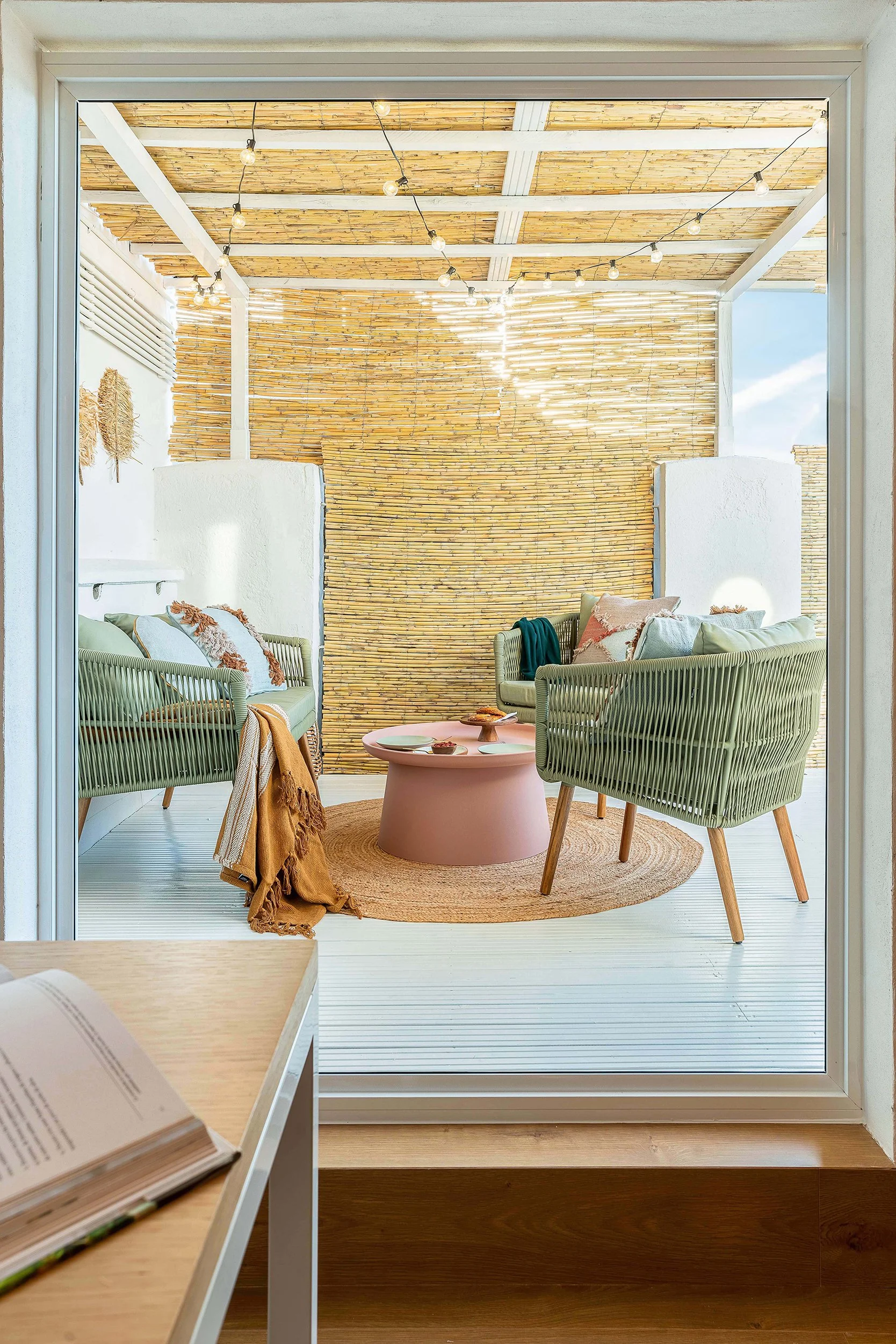 Cozy patio space with rattan furniture, a pink coffee table, and a woven rug, enclosed by bamboo walls and a pergola adorned with string lights.