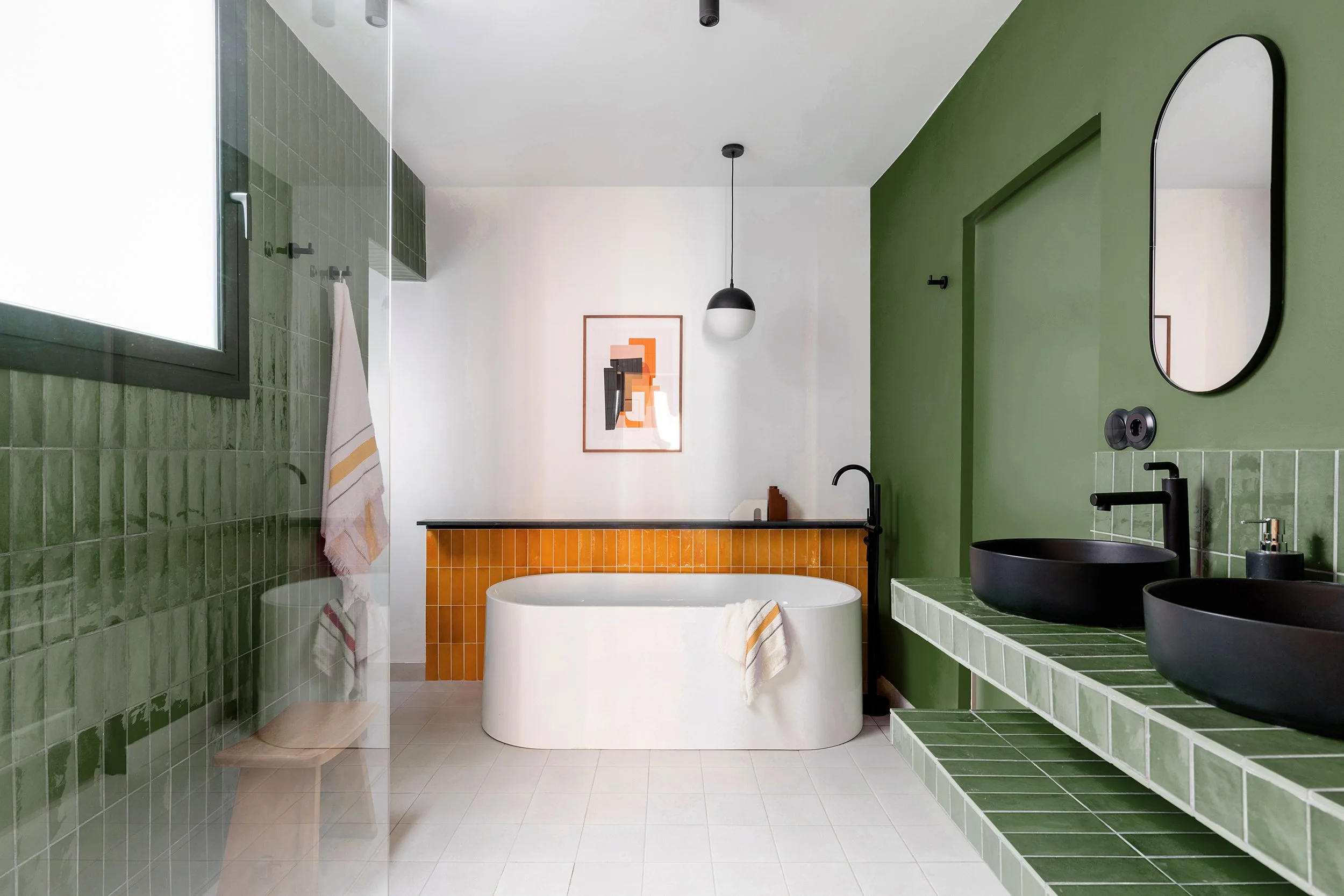 Modern bathroom with green tile walls, a white freestanding bathtub, black faucets, twin black vessel sinks, a round hanging light, and an abstract artwork.