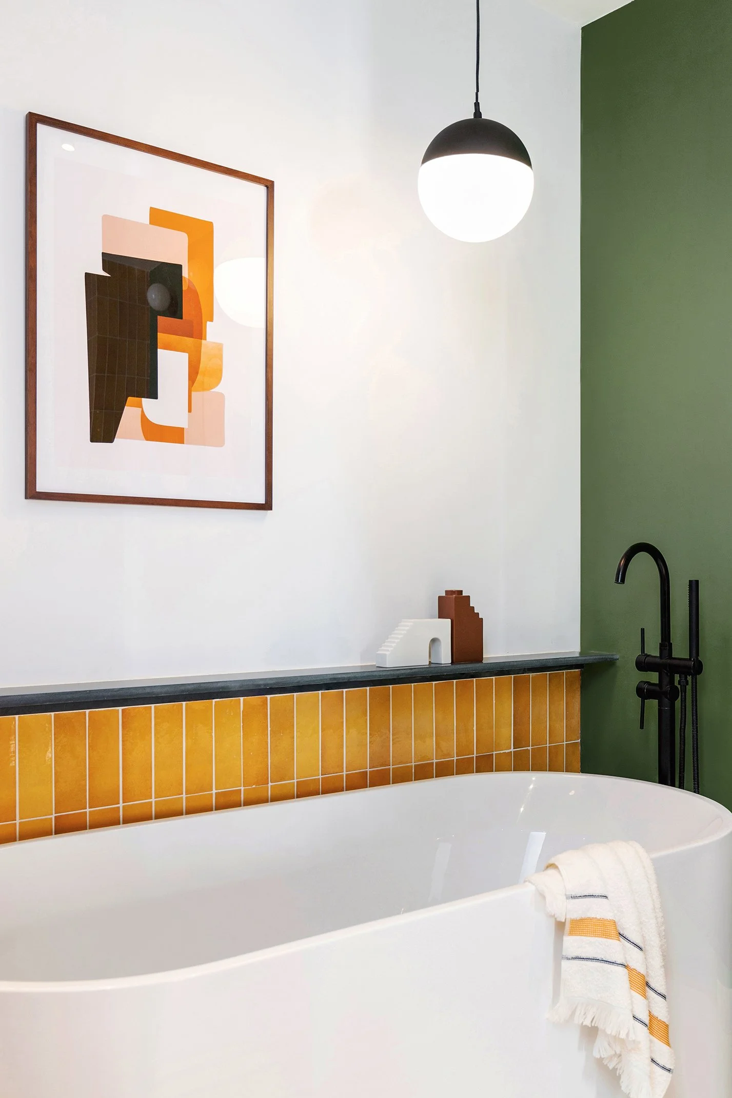 Modern bathroom with white bathtub, yellow tiled wall, green accent wall, black faucet, hanging pendant light, and abstract art.