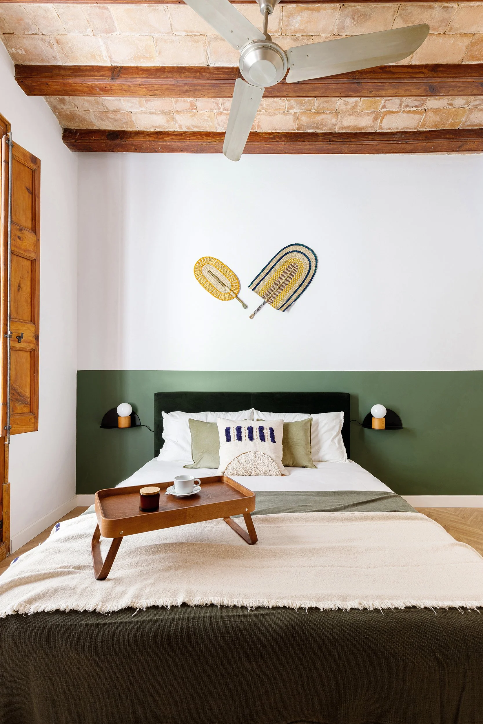 Cozy bedroom with a bed, green and white bedding, wooden tray with cup, ceiling fan, wall decor, and rustic wooden beams on the ceiling.
