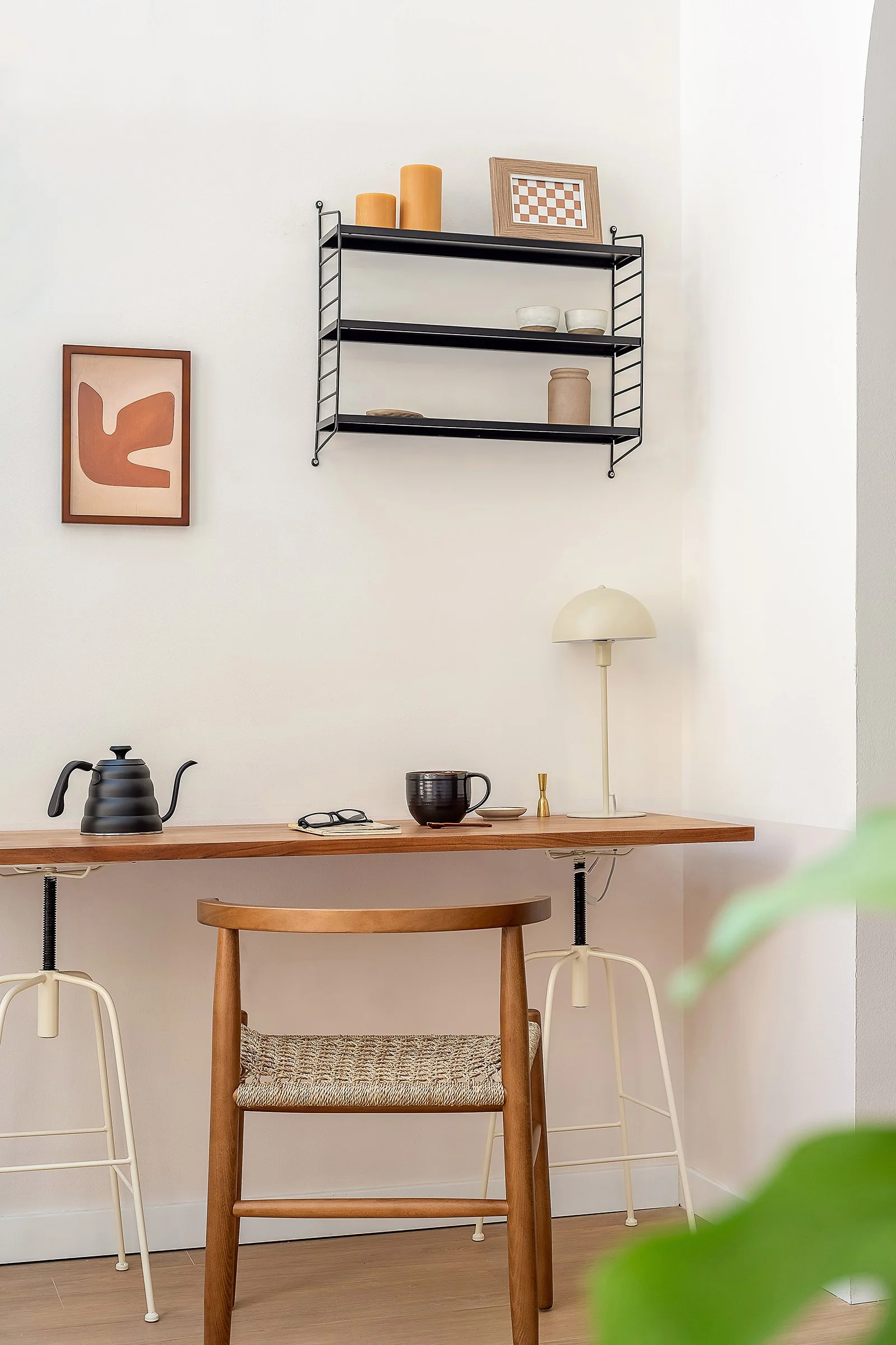 Minimalist home office with wooden desk, chair, and shelves; includes black kettle, mug, lamp, framed art, and decorative items.