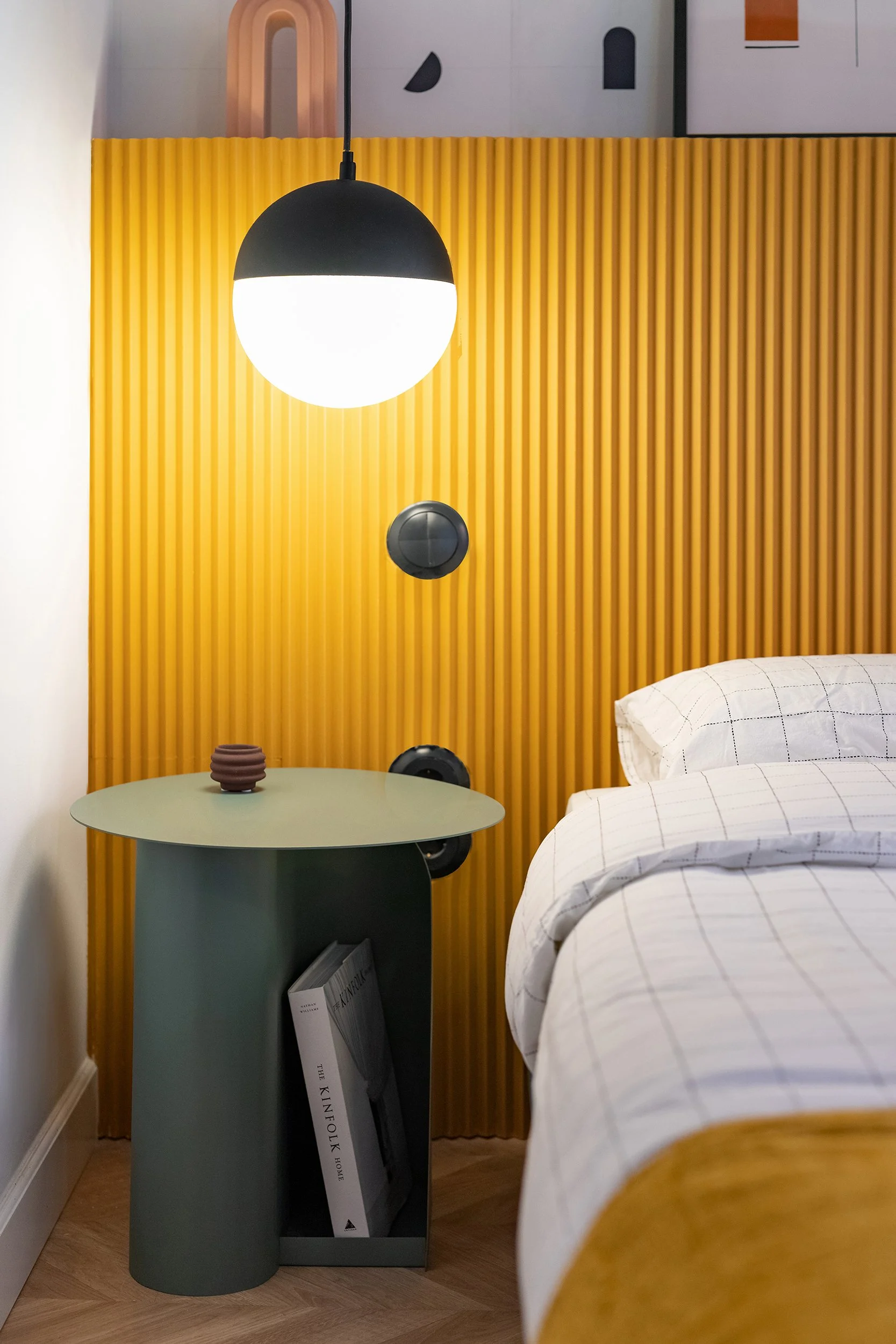 Modern bedroom corner with a round hanging lamp, yellow corrugated wall panel, green side table, book, and plaid bedding.