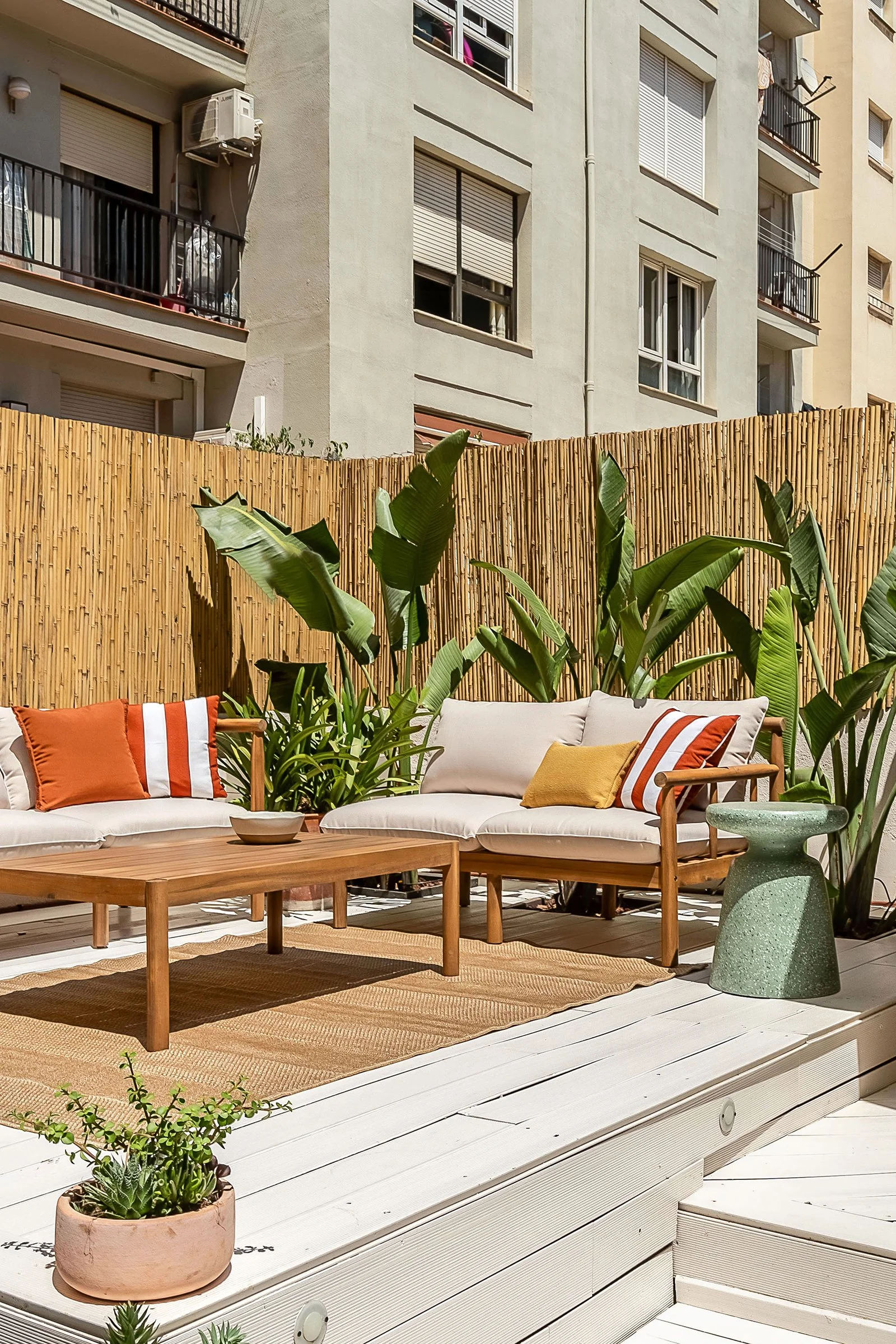 Outdoor patio with wooden furniture, cushions, a bamboo privacy fence, and potted plants, surrounded by apartment buildings.
