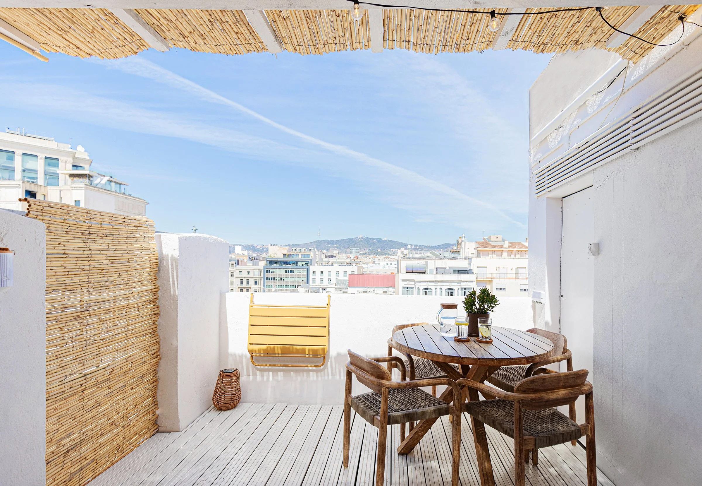 Rooftop terrace with wooden furniture, bamboo privacy screen, potted plants, and cityscape view.