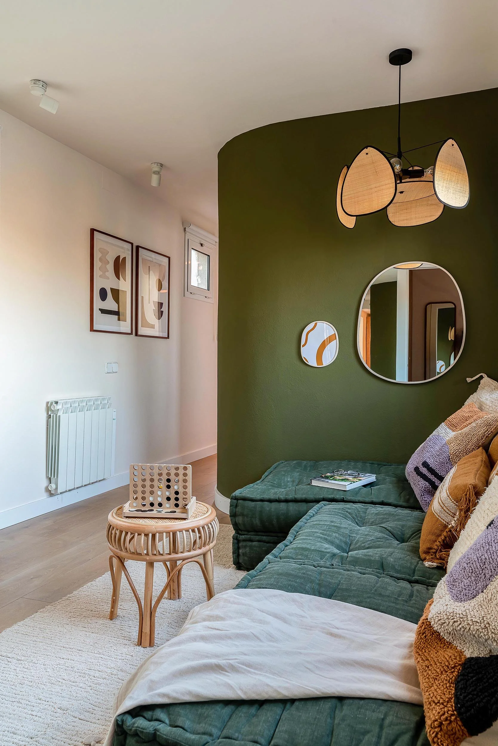 Modern living room with olive green accent wall, decorative pillows on a green sofa, round wall mirrors, abstract framed art, wicker side table, game board, pendant light, and hardwood floor.