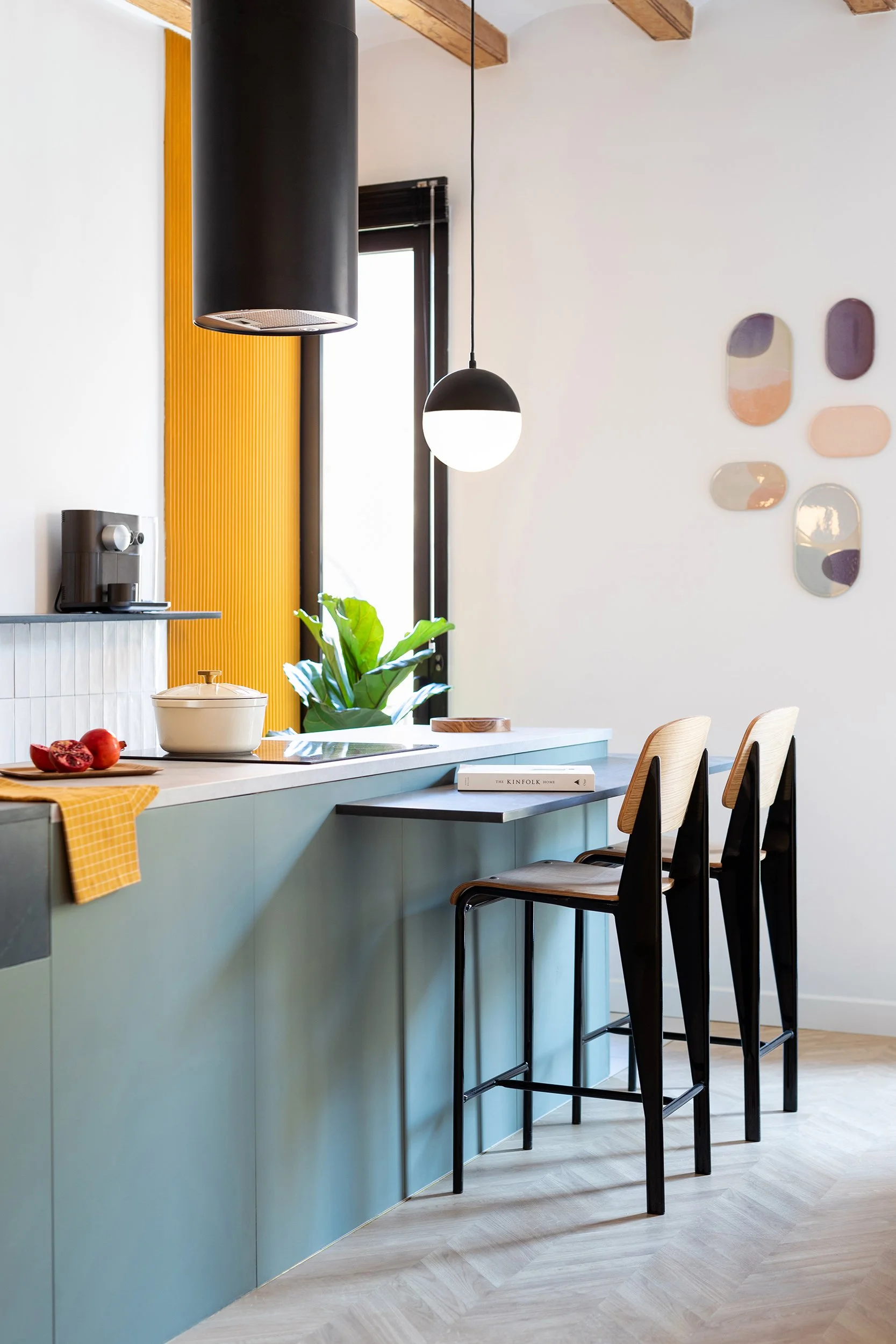Modern kitchen interior with blue cabinetry, black pendant lights, a countertop, potted plant, and barstools. A pomegranate and yellow cloth are on the counter, with wall art and a book on the side.