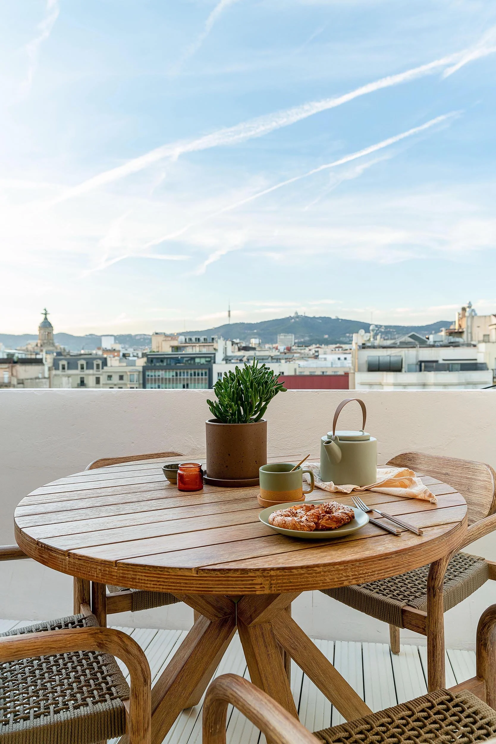 Rooftop patio with round wooden table, a plant, food and drinks, cityscape and mountain view.