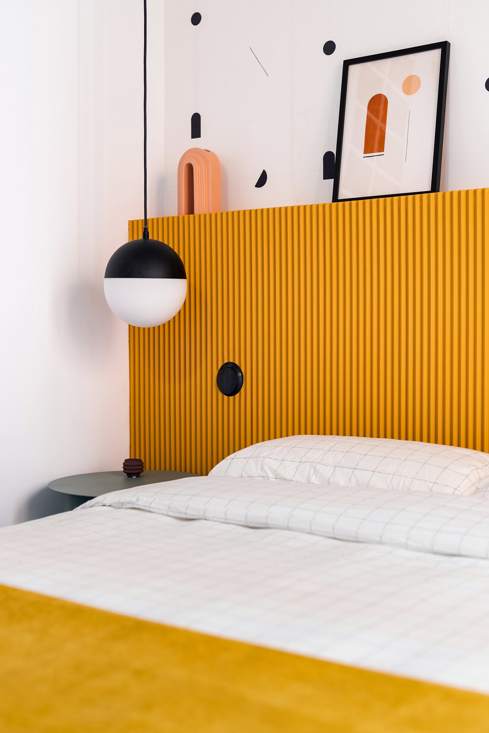 Modern bedroom with a yellow ribbed headboard, white and black spherical pendant light, grid-patterned bedding, and abstract art on the wall.