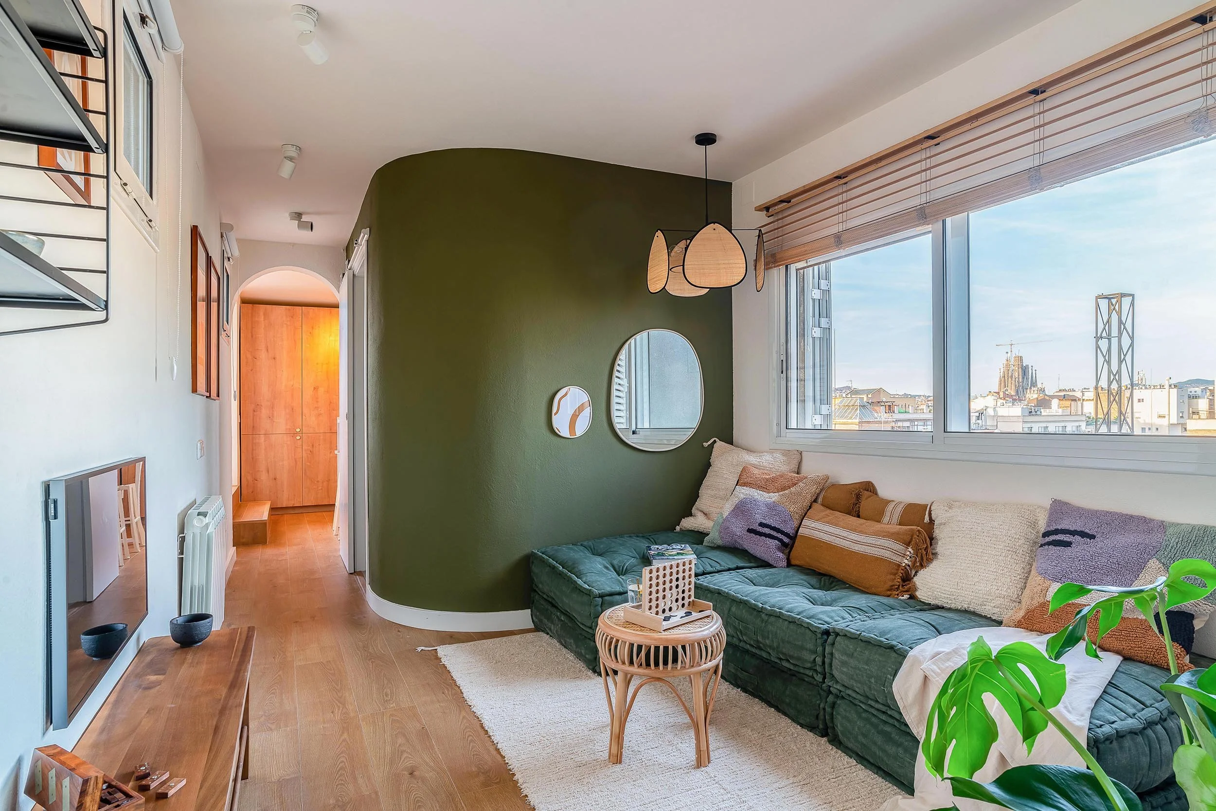 Modern living room with green L-shaped sectional sofa, round rattan coffee table, and decorative pillows. The room features a curved green accent wall, mirrors, and pendant lights. Large window with cityscape view and wooden blinds. Wooden flooring a
