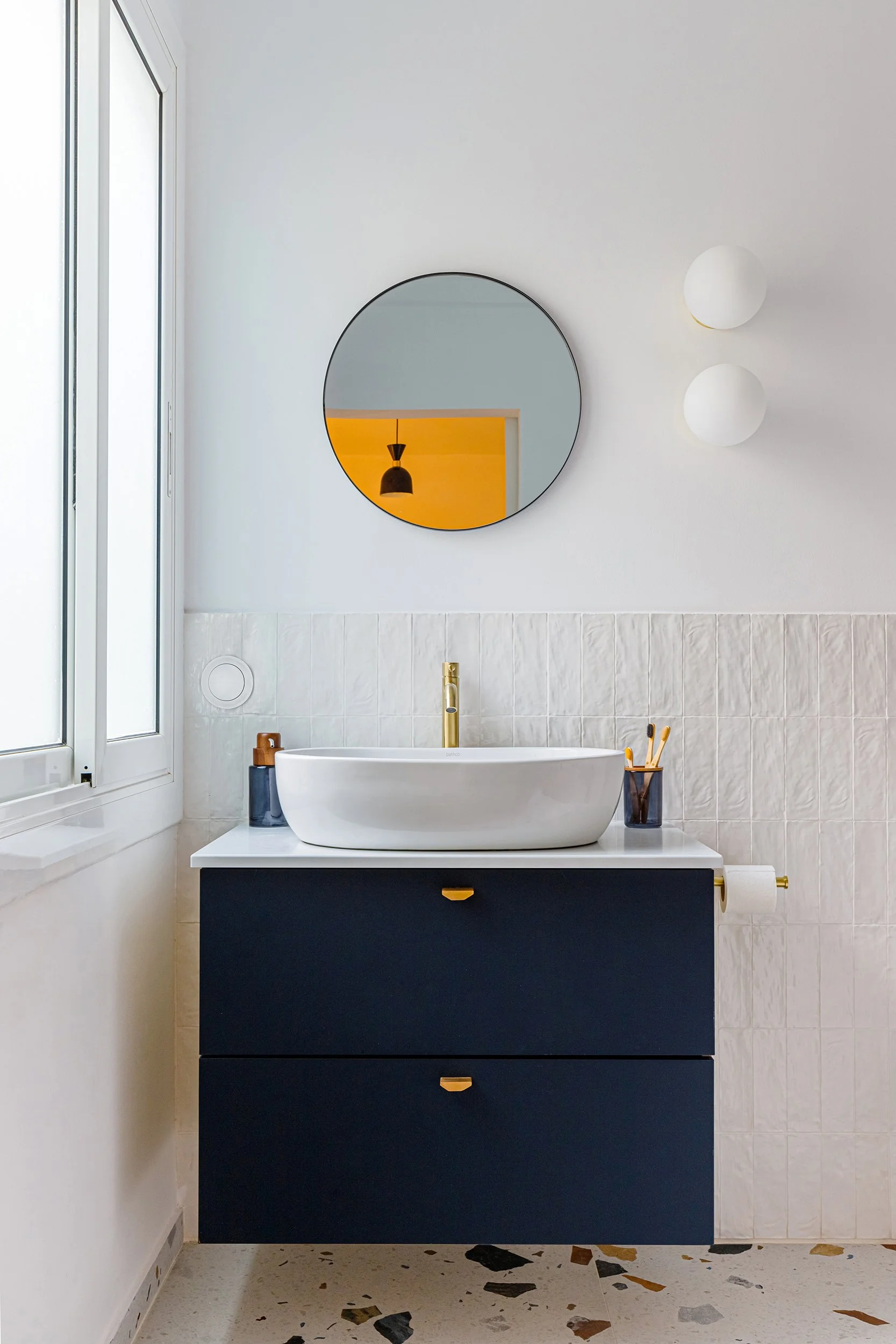 Modern bathroom with round mirror, white sink, gold faucet, navy blue vanity, tiled wall, and terrazzo floor.