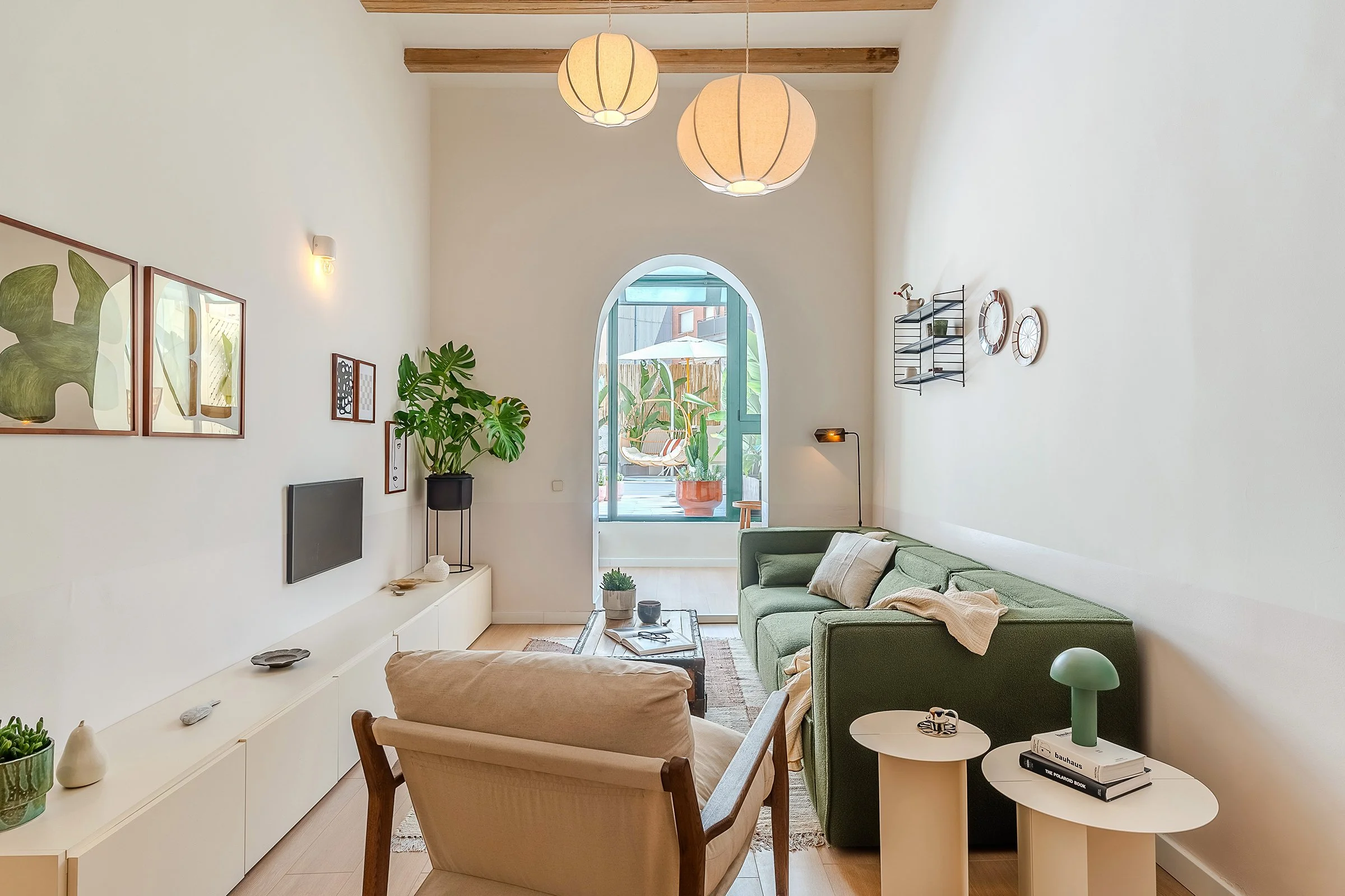 Contemporary living room with a green sofa, armchair, wall lamps, a white cabinet, indoor plants, and decorative art. Features a large arched window leading to an outdoor space with plants and a sitting area.