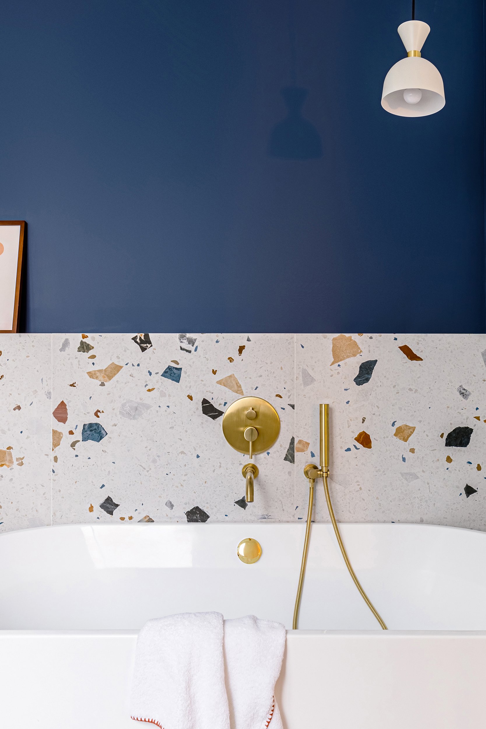 Modern bathroom with a white bathtub, terrazzo tiles, brass fixtures, a hanging light fixture, and a towel on the edge of the tub against a blue wall.