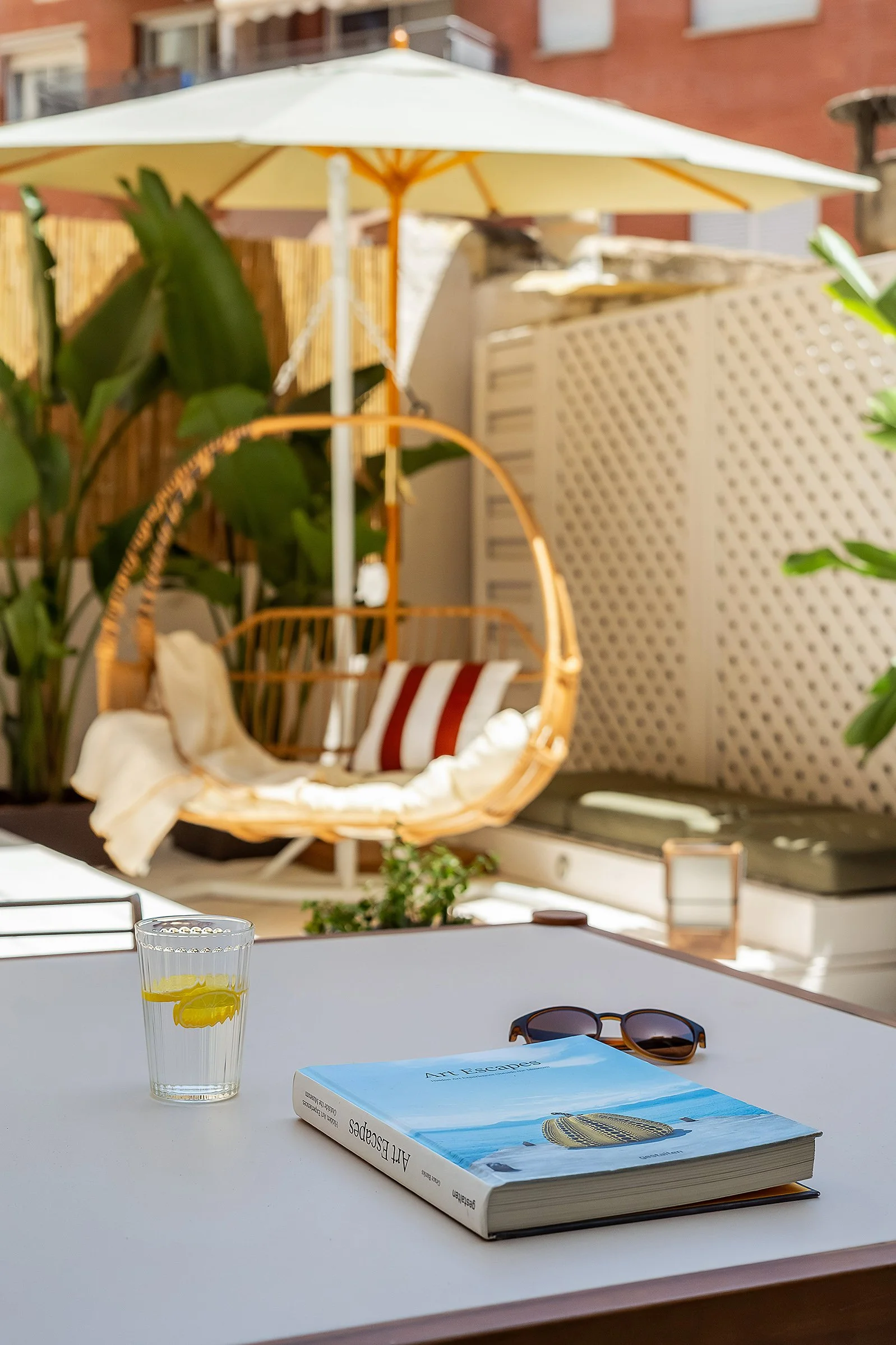 Outdoor patio with a book titled 'Art Escapes,' sunglasses, and a glass of water with lemon on a table. In the background, a hanging chair with cushions under an umbrella, surrounded by plants.