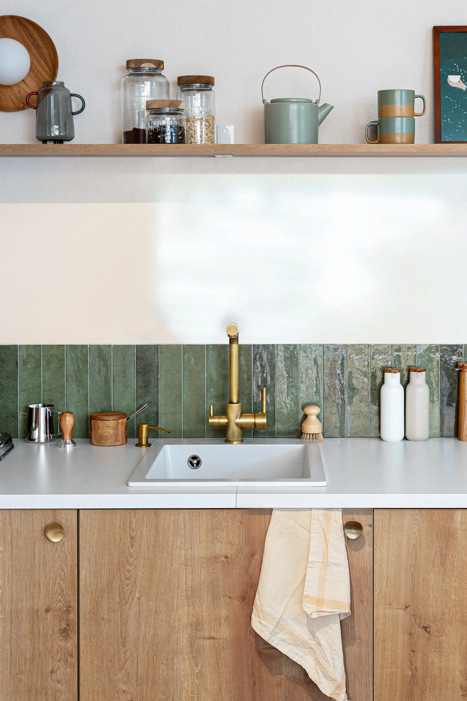 Modern kitchen with wooden cabinets, white countertop, green tile backsplash, brass faucet, and shelf with jars, kettle, and mug.