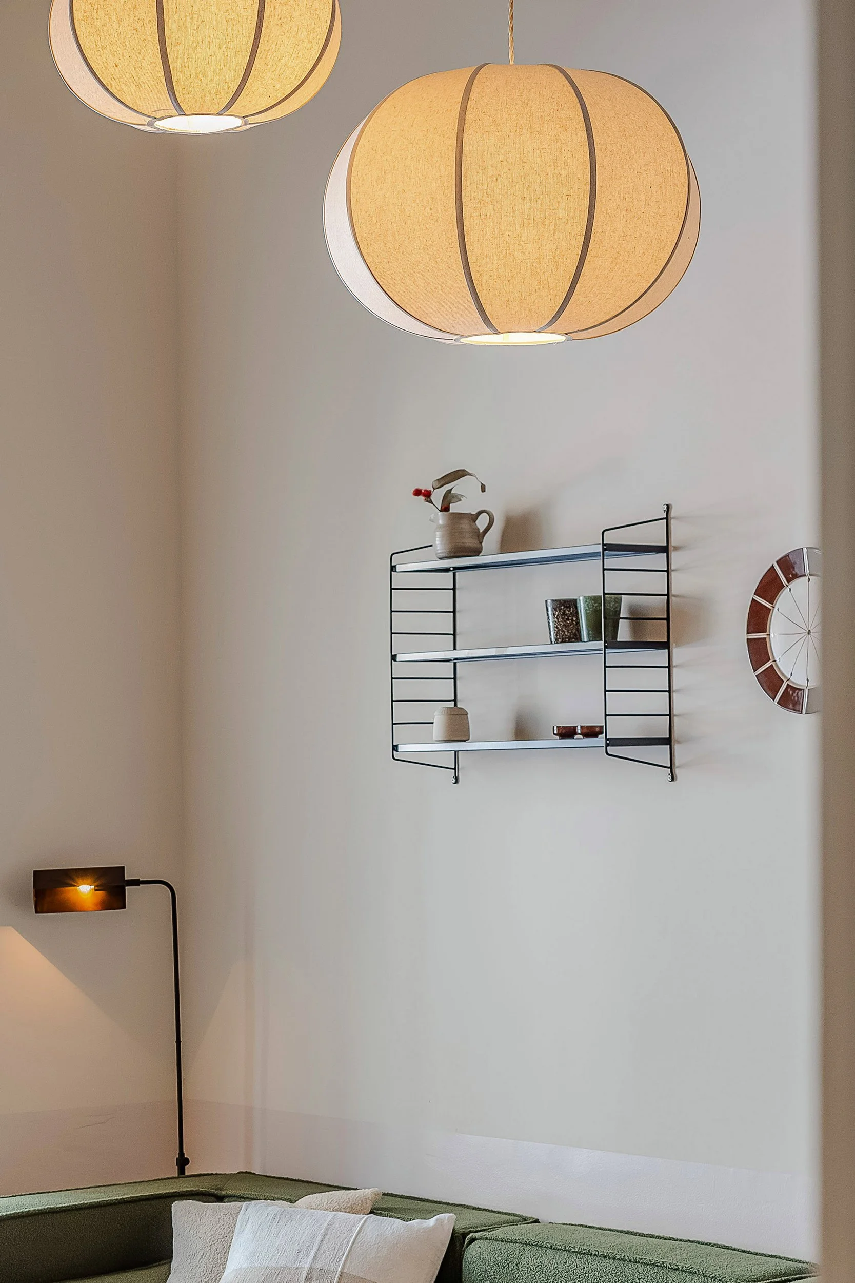 Interior room with two hanging spherical pendant lights, a black wall-mounted shelf with decorative items, a wall sconce, and a green couch with a white pillow.