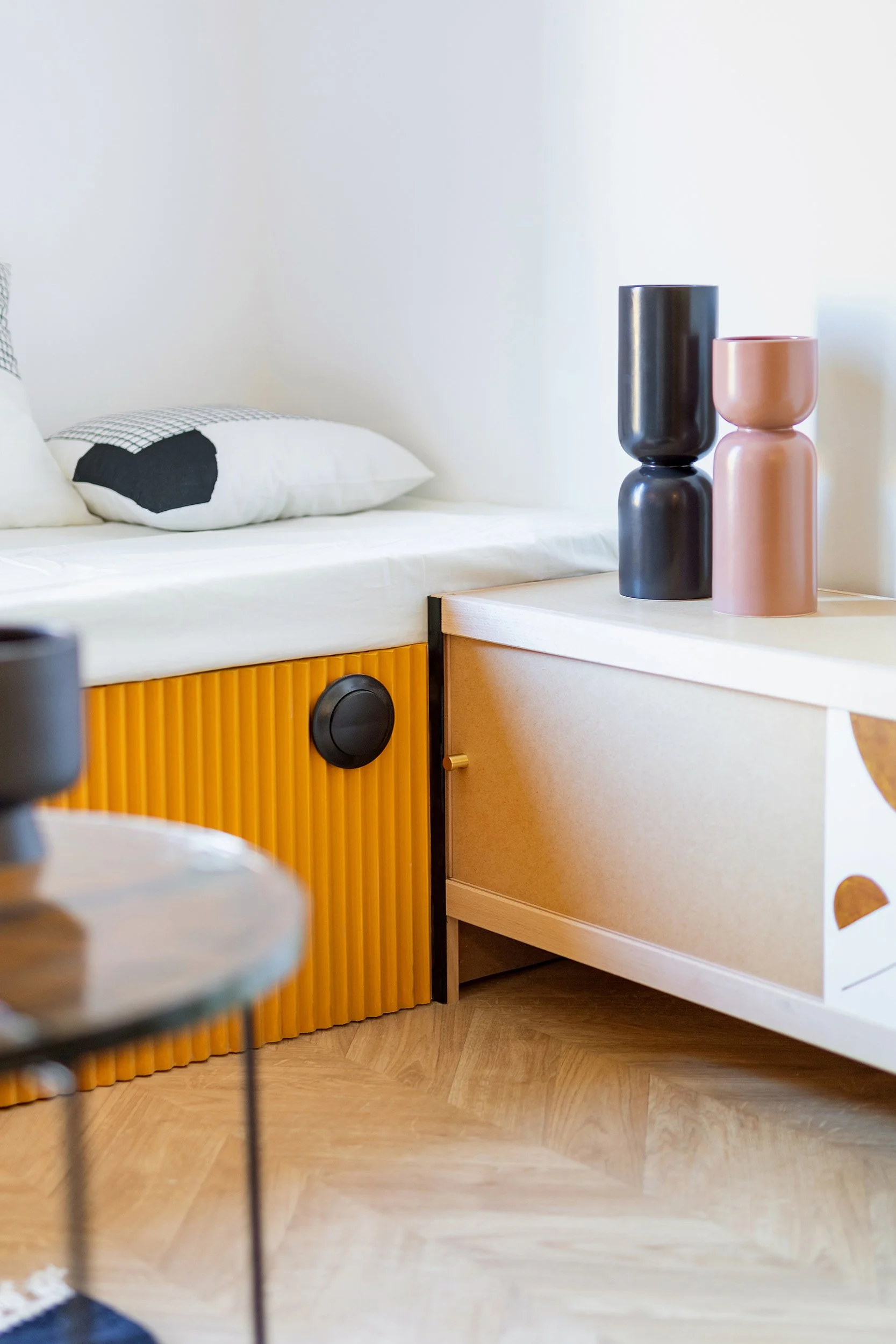 Modern interior design featuring a white cushion on a bench with a black and white patterned pillow, a yellow ribbed storage unit, two ceramic vases in black and brown tones on a wooden cabinet, and a glass-topped side table on a wooden floor.