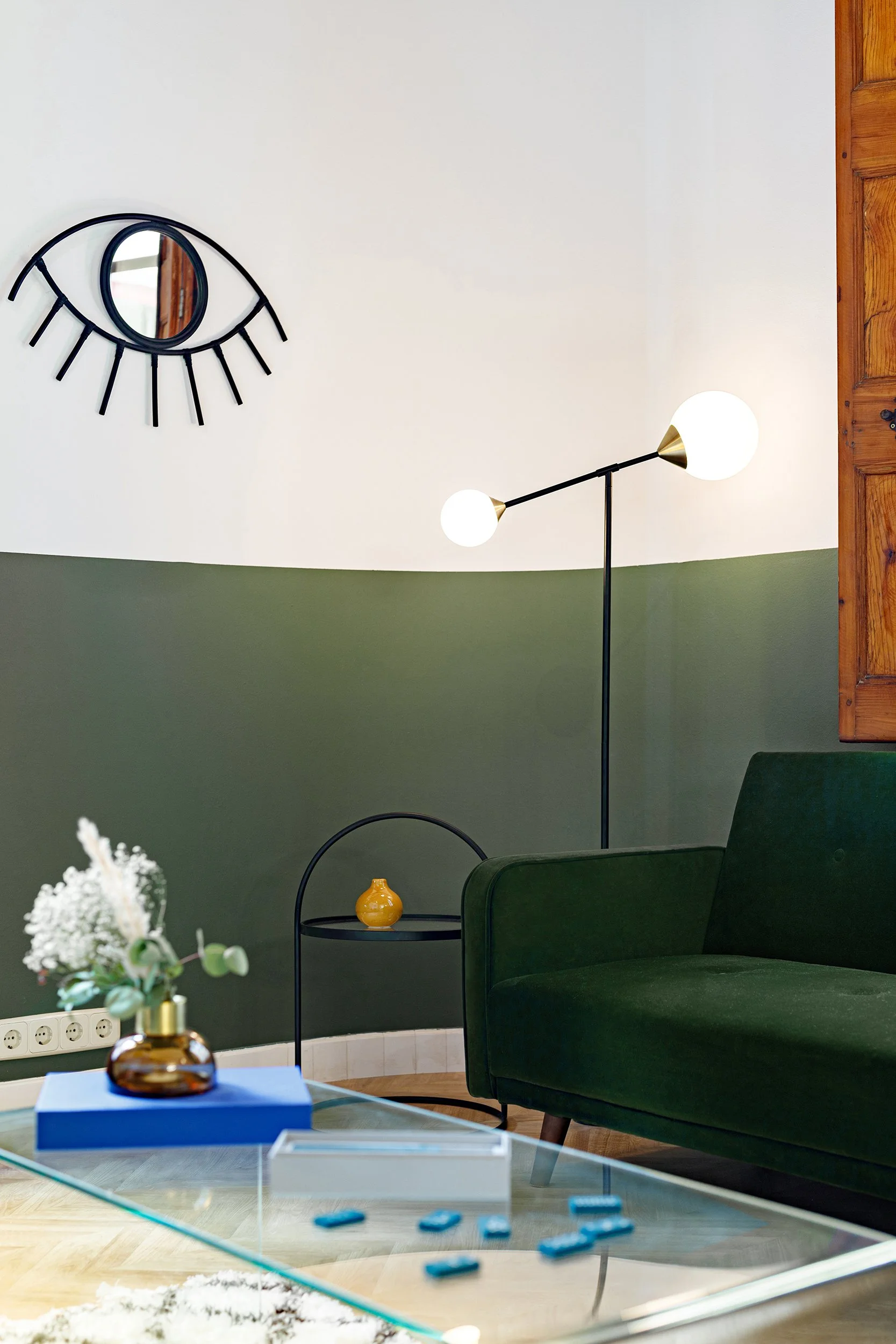 Modern living room with green velvet sofa, glass table, decorative wall mirror shaped like an eye, two-globe floor lamp, small side table with a yellow vase, and white flowers in a brown vase.