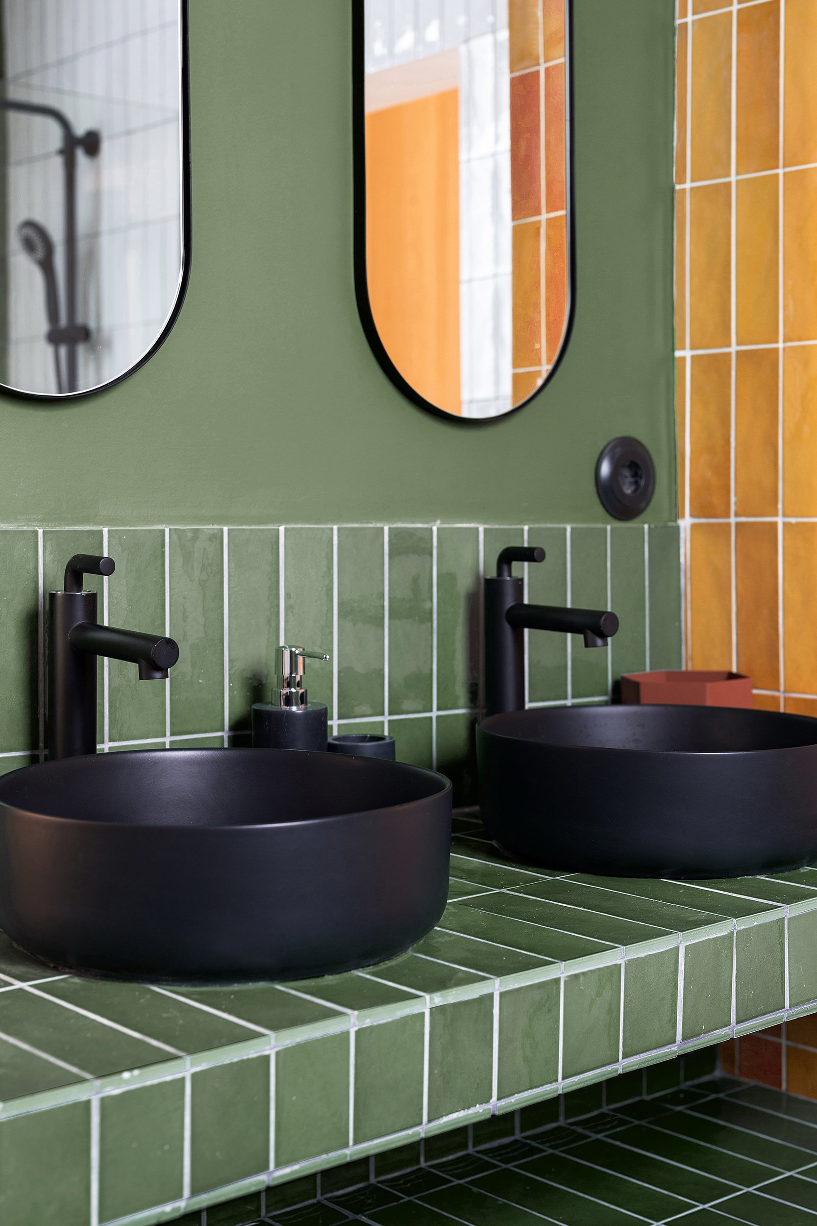 Modern bathroom with two black vessel sinks and matte black faucets on a green tiled countertop, green and orange wall tiles, and vertical oval mirrors.