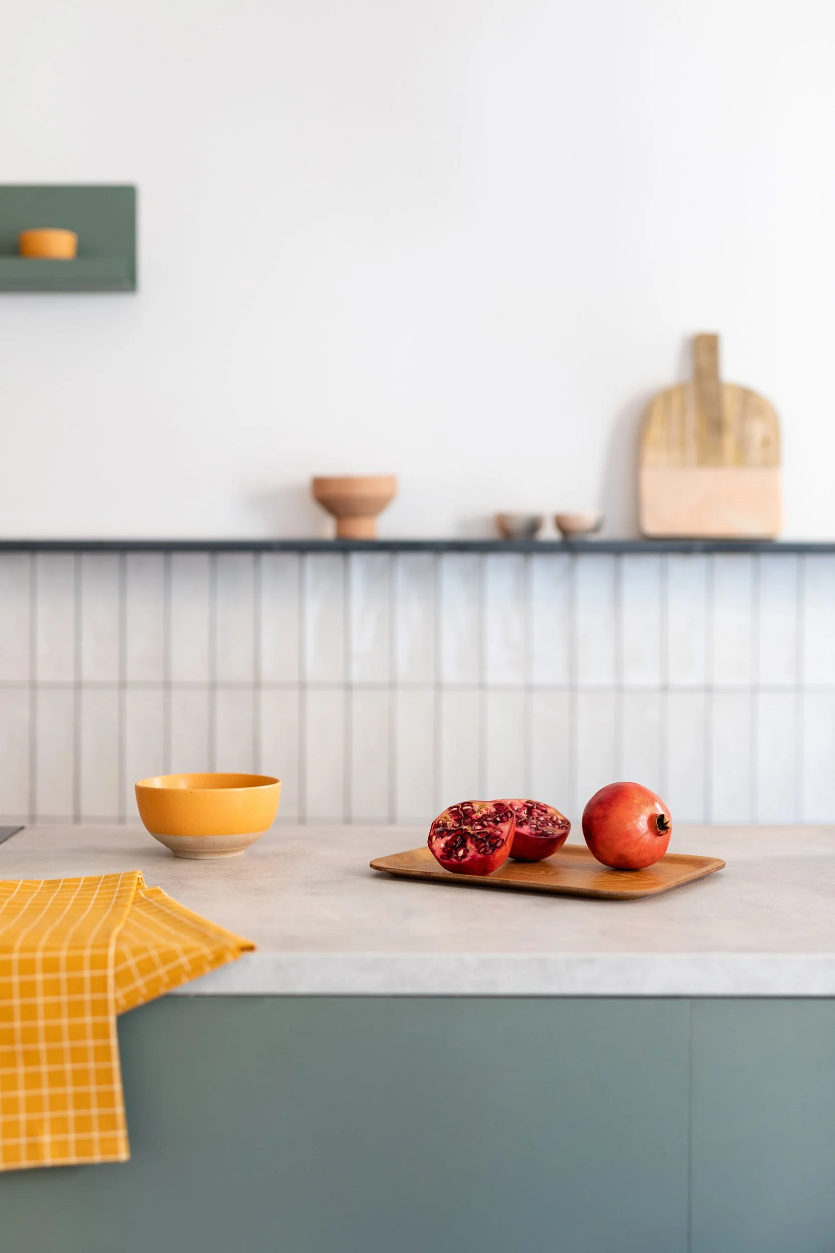 Kitchen countertop with a whole and halved pomegranate on a wooden tray, yellow bowl, and checkered cloth in a modern kitchen setting.