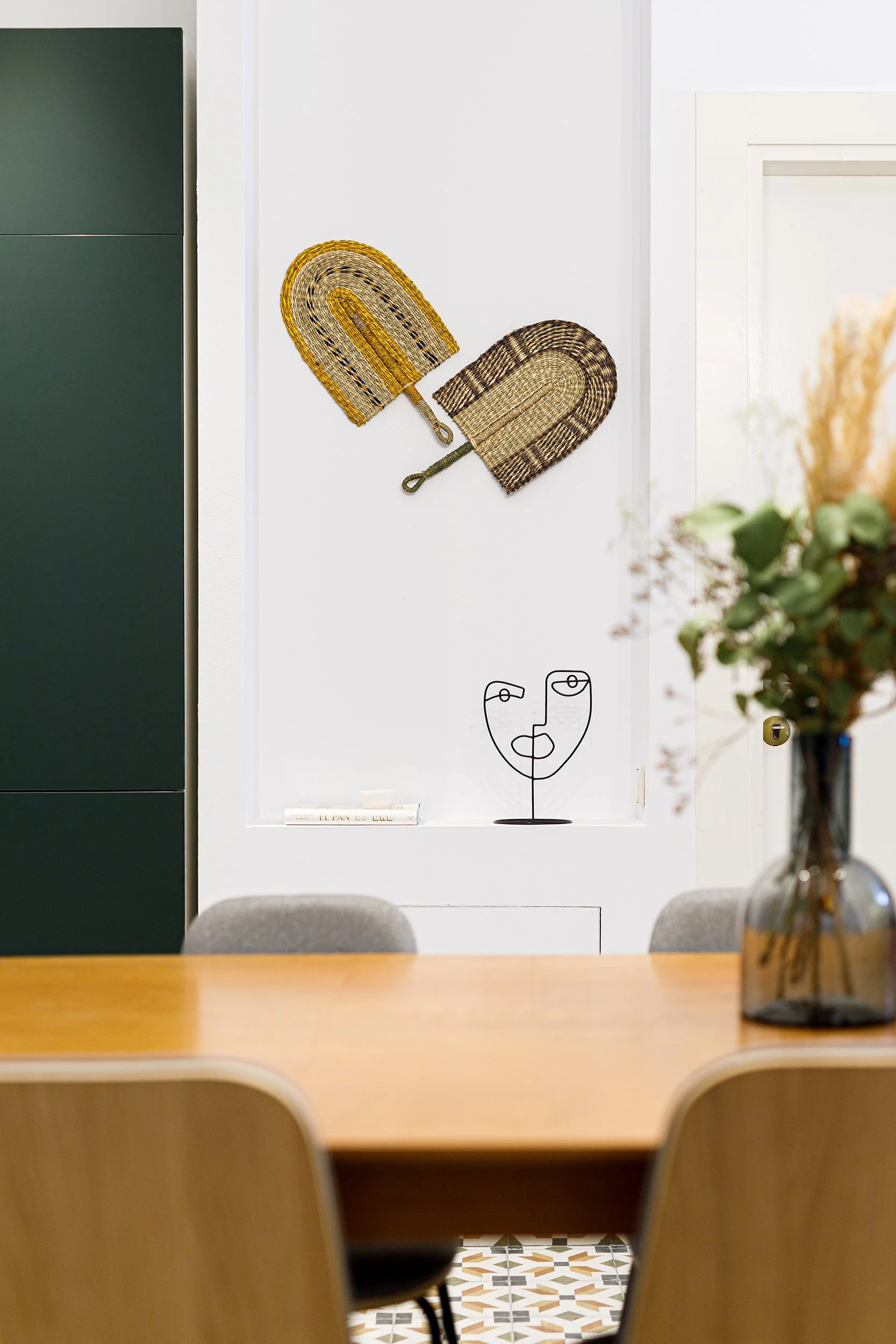 Interior of a dining area with a wooden table, modern chairs, a vase with greenery, two woven fans on the wall, and minimalist art on a shelf.