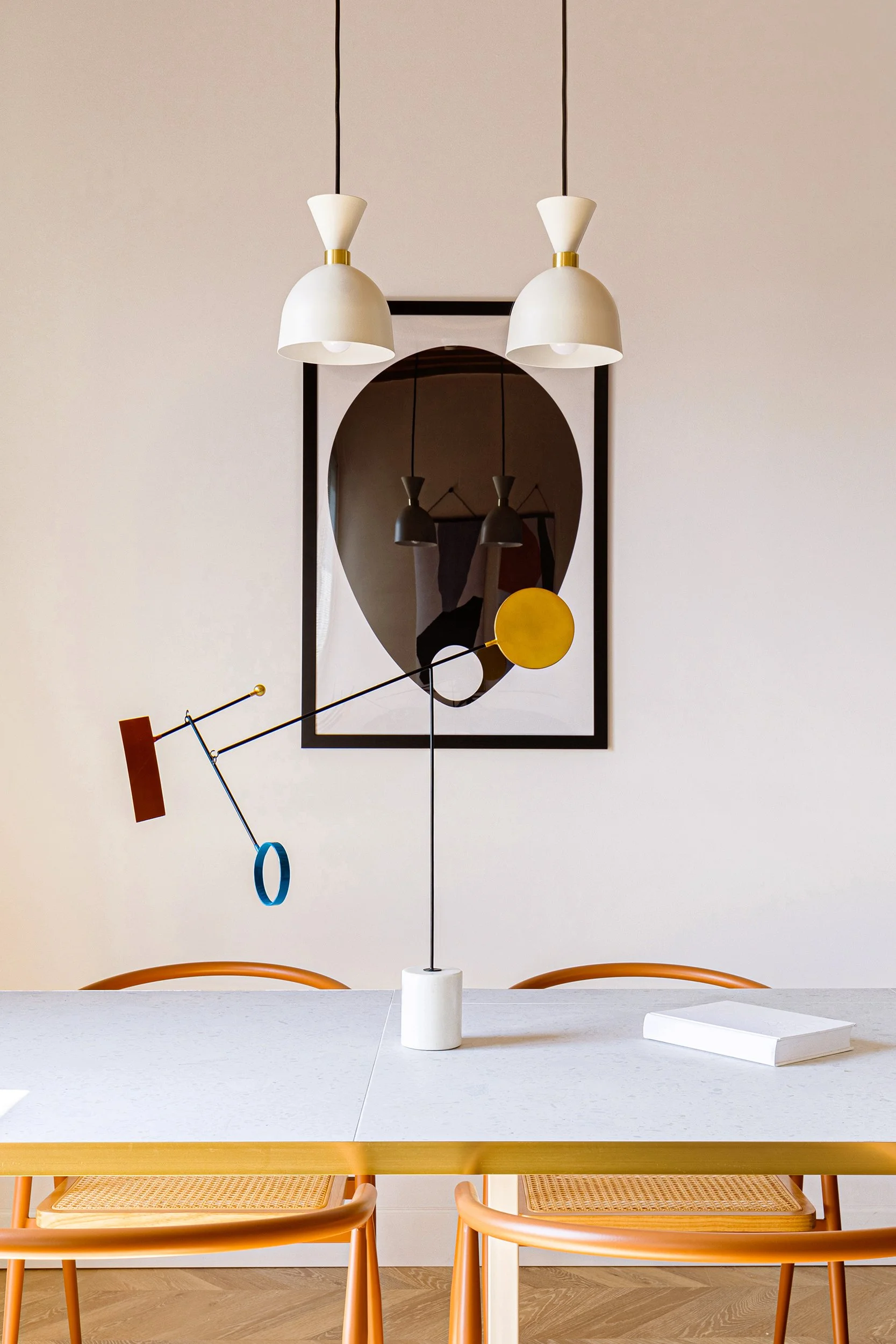 Modern dining room with orange chairs and white table, decorated with a mobile sculpture and pendant lights, and featuring abstract artwork on the wall.