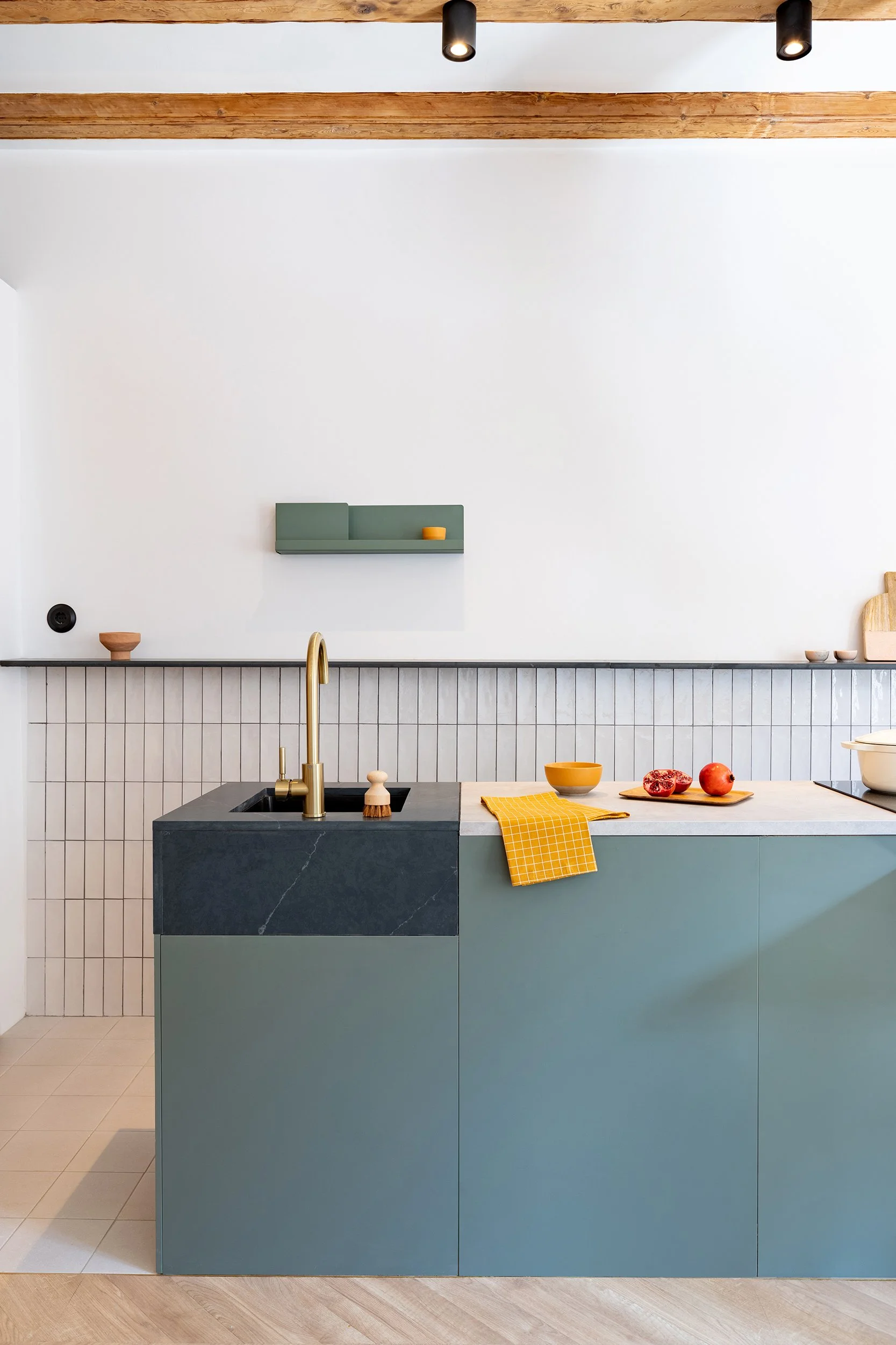 Modern kitchen with teal cabinets, a black countertop, and gold faucet; features a tiled backsplash and wooden ceiling beams. Kitchen items include a yellow towel, a brush, a bowl, and fruit.