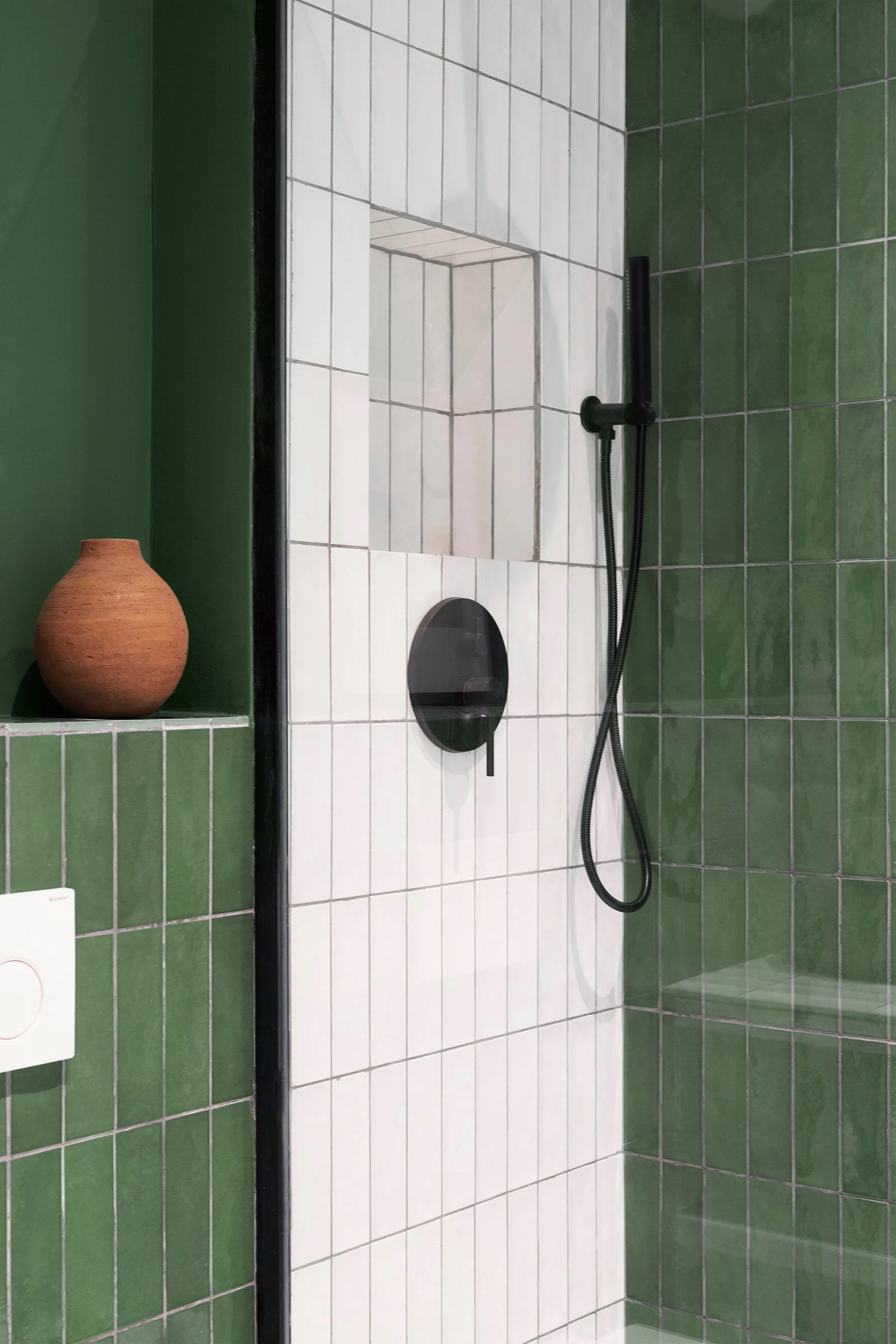 Modern bathroom with green and white tiled walls, black shower fixture, and a terracotta vase on a shelf.