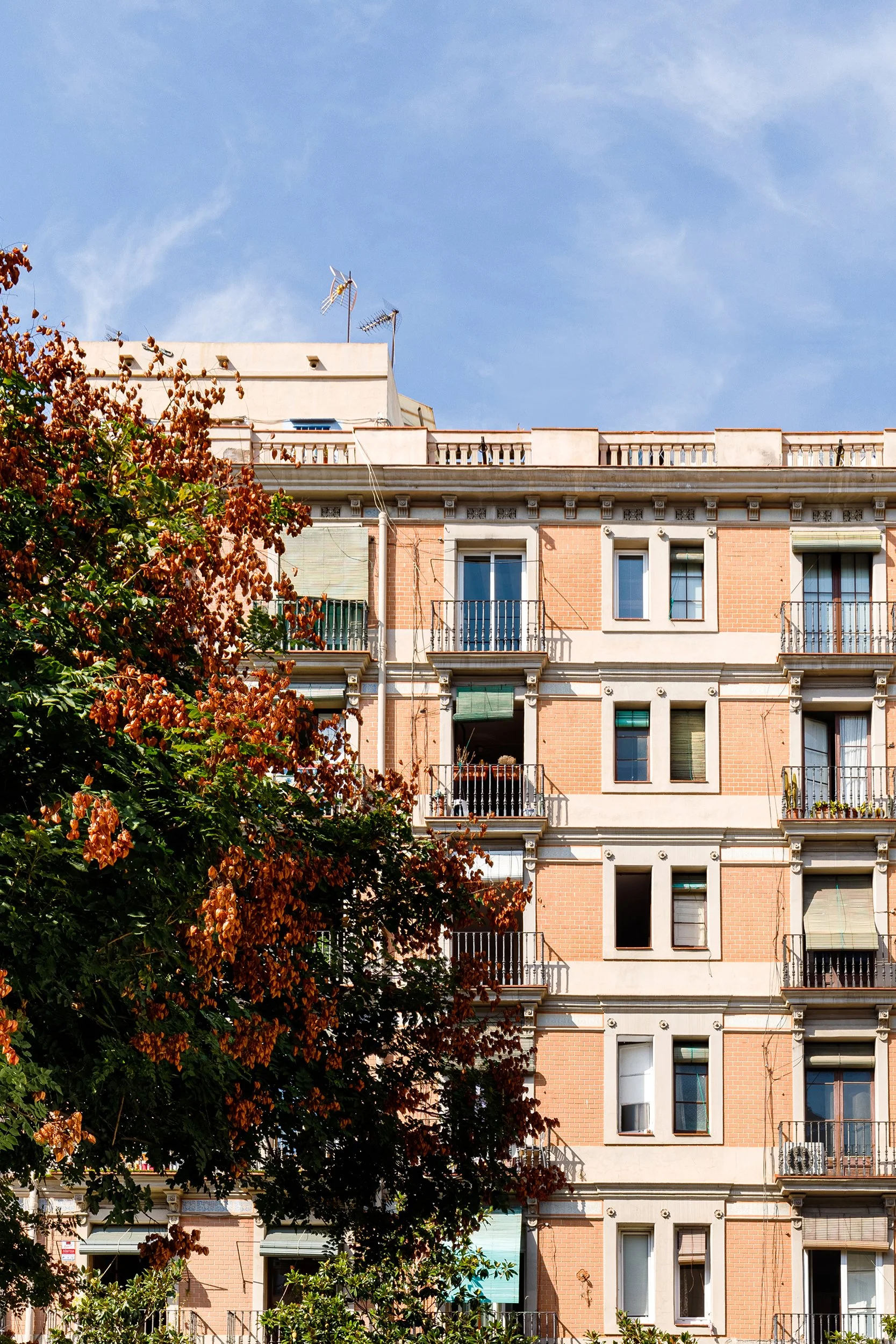 Exterior of an apartment building with balconies, surrounded by trees with orange leaves, under a blue sky.