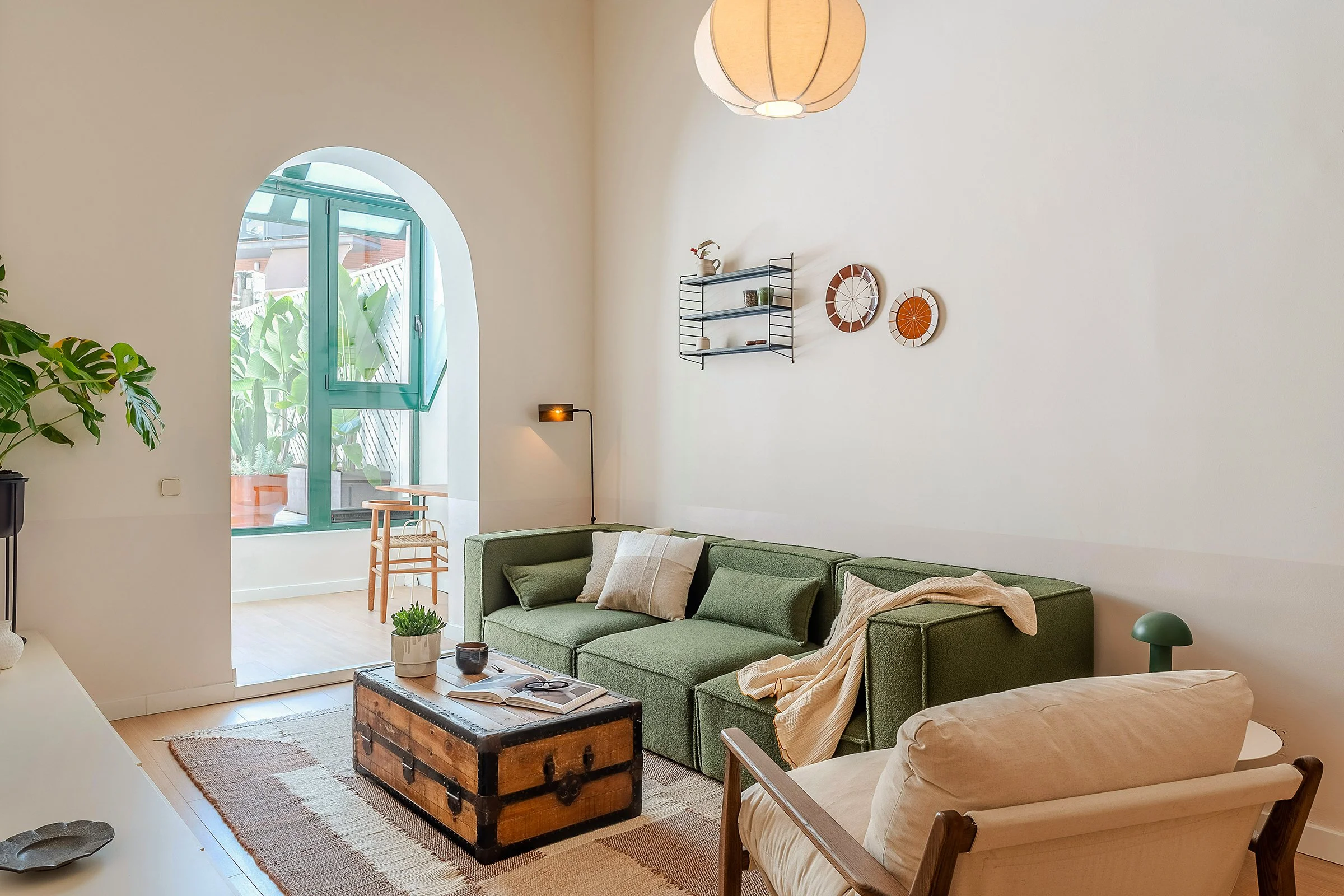 Cozy living room with green sectional sofa, wooden coffee table, and indoor plants. Features beige walls, wall-mounted shelves, and a large arched window leading to a terrace. Bohemian decor style with natural light and a cream-colored armchair.
