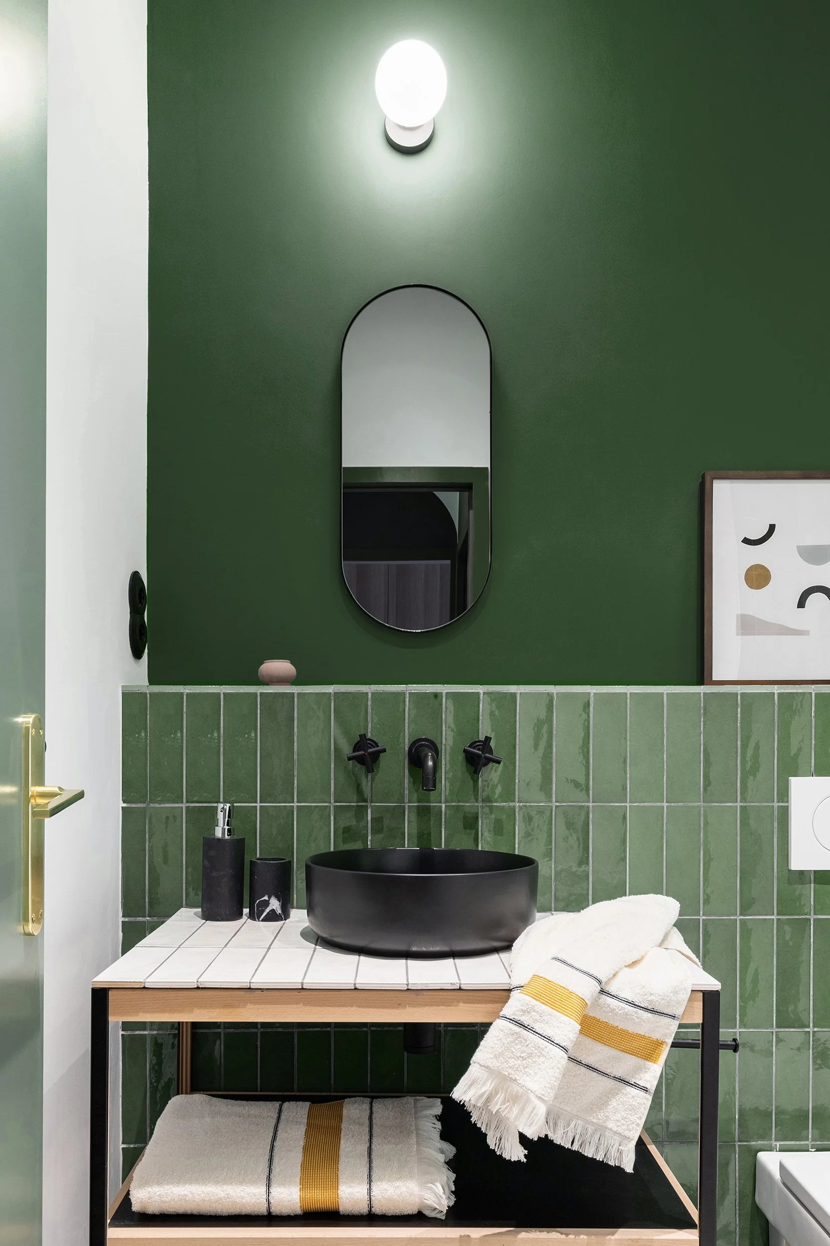 Modern bathroom with green walls and matching green tiled backsplash, featuring a black sink, black fixtures, a circular mirror, and a light fixture above. White and yellow-striped towels are neatly arranged. Minimalist decor includes a simple framed